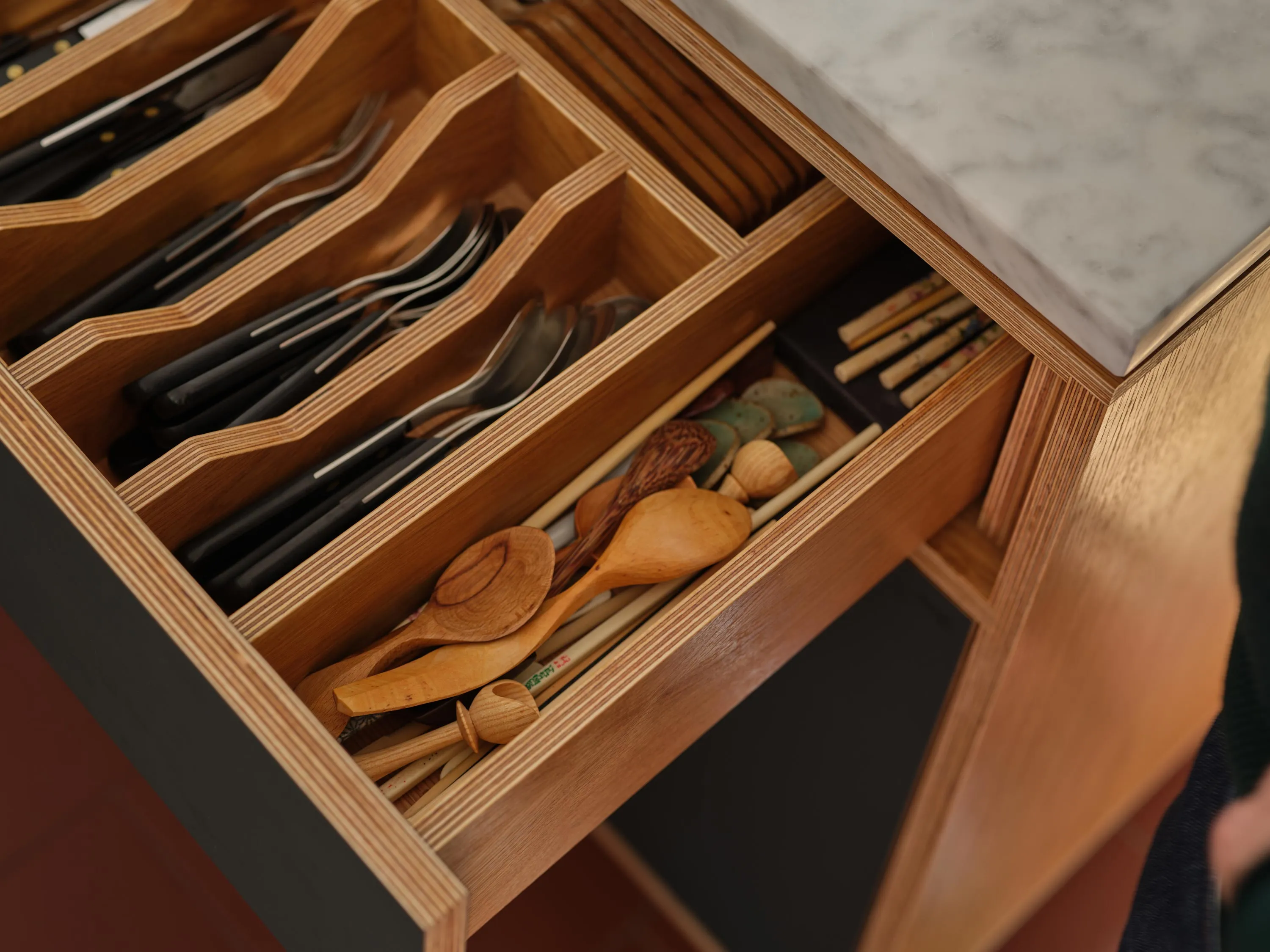 Open kitchen drawer with organized black-handled knives, forks, spoons, wooden spoons, chopsticks, and ceramic chopstick rests.