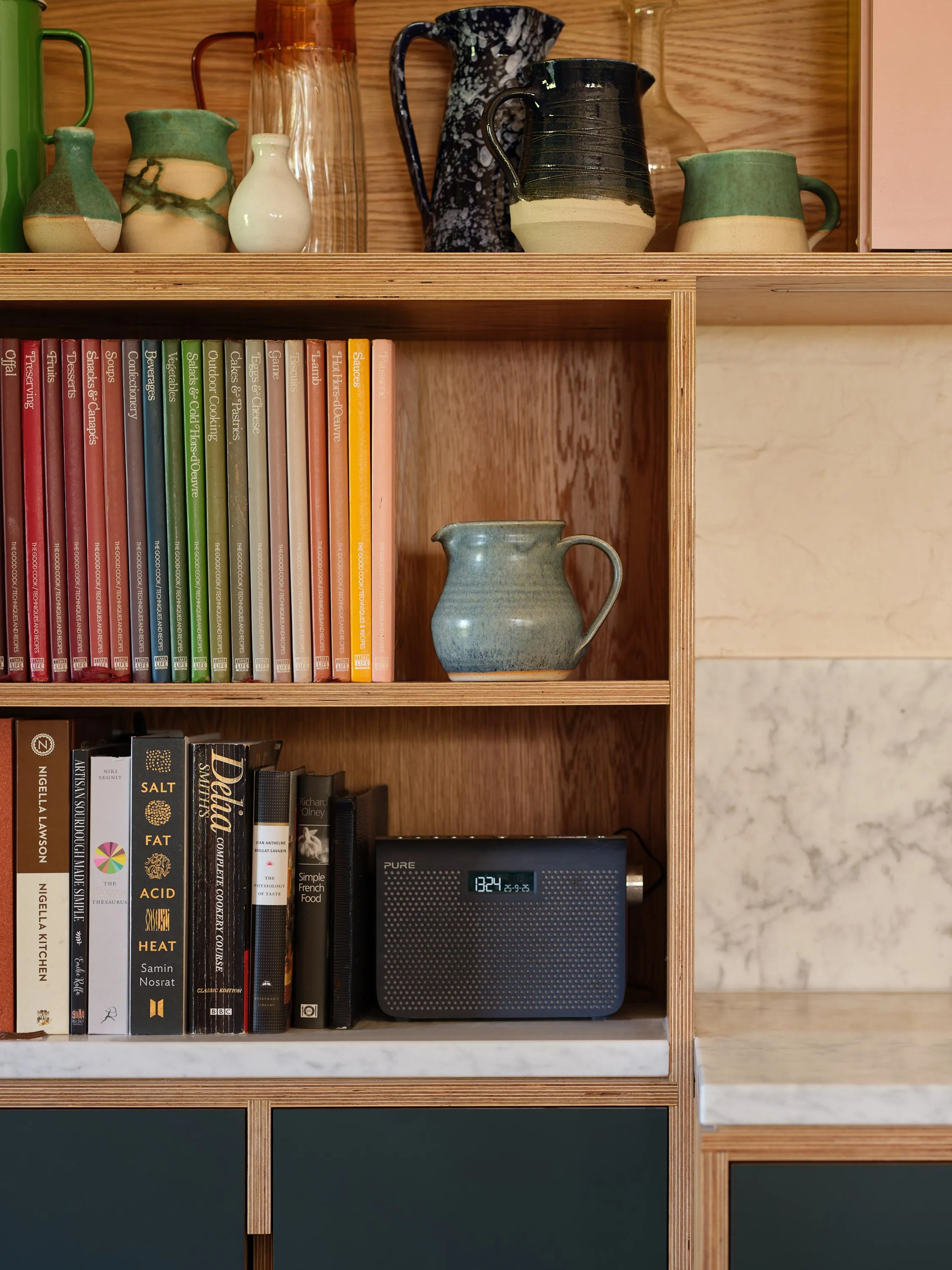Wooden shelves with cookbooks, ceramic pitchers, and a digital radio displaying time 13:24.
