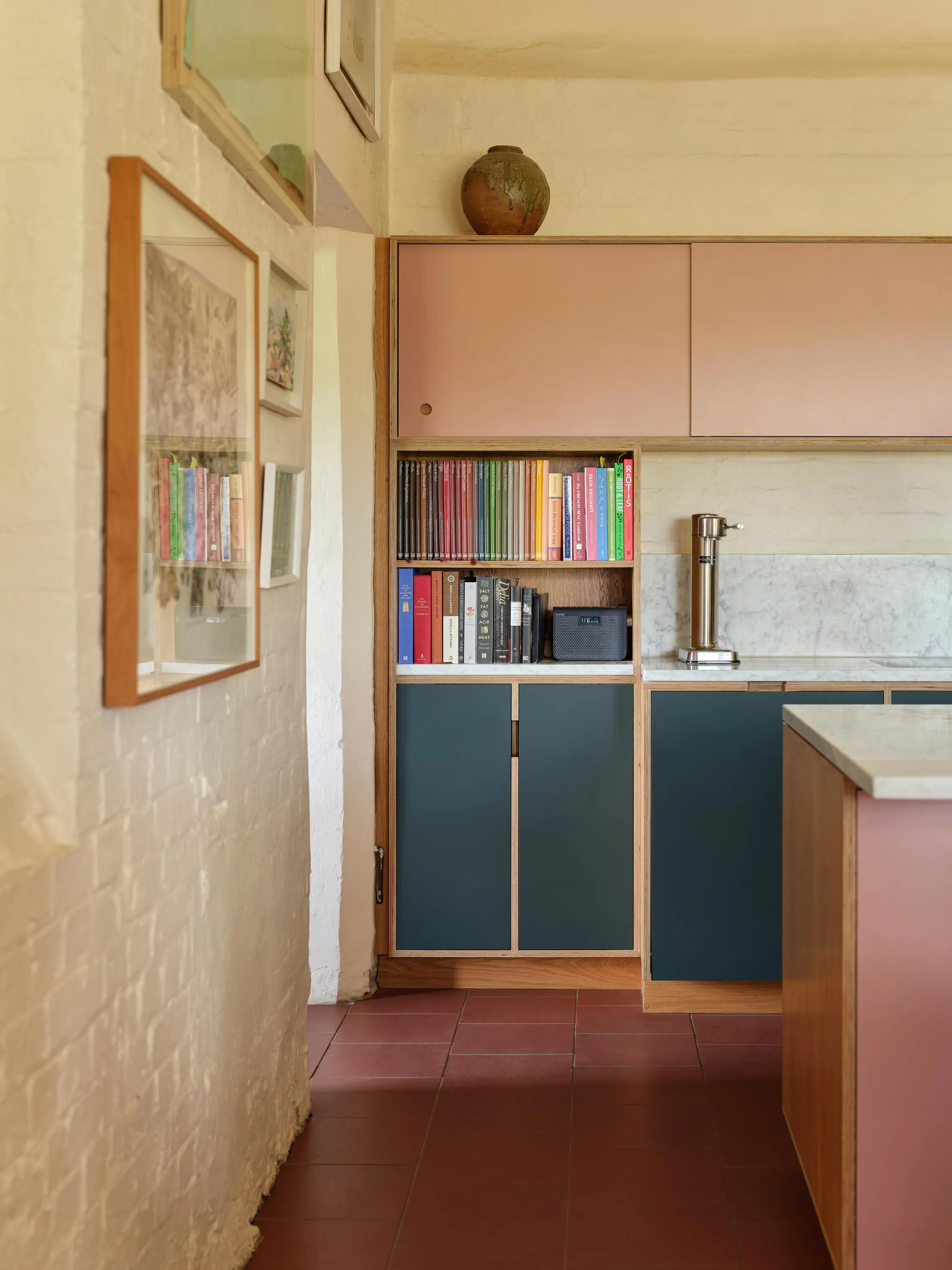 Kitchen with pink upper cabinets, teal lower cabinets, a marble backsplash, a countertop with a brass soda siphon, and a bookshelf with colorful books and a speaker.