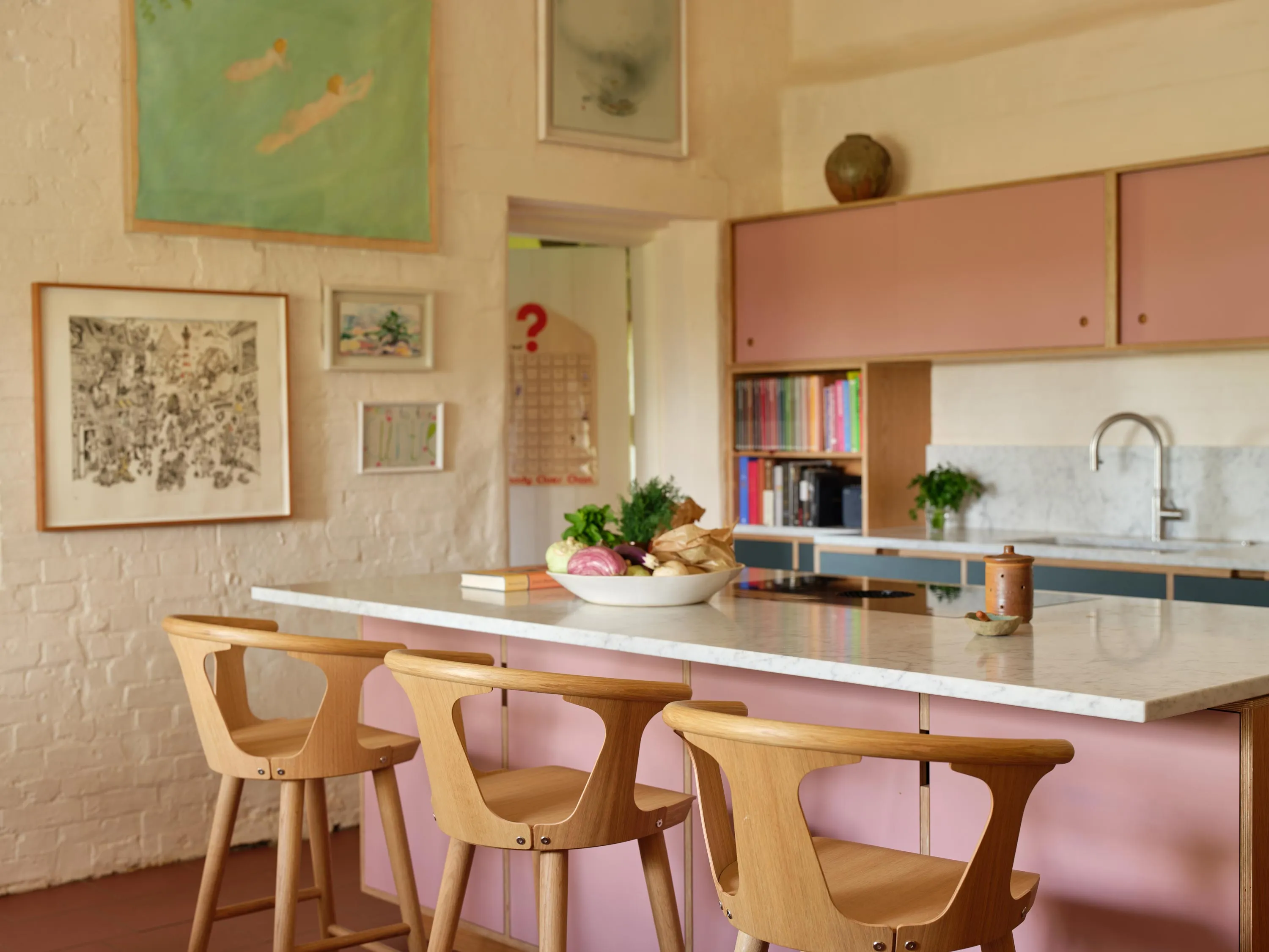 Modern kitchen with pink cabinetry, wooden bar stools, marble countertop island, and wall art.