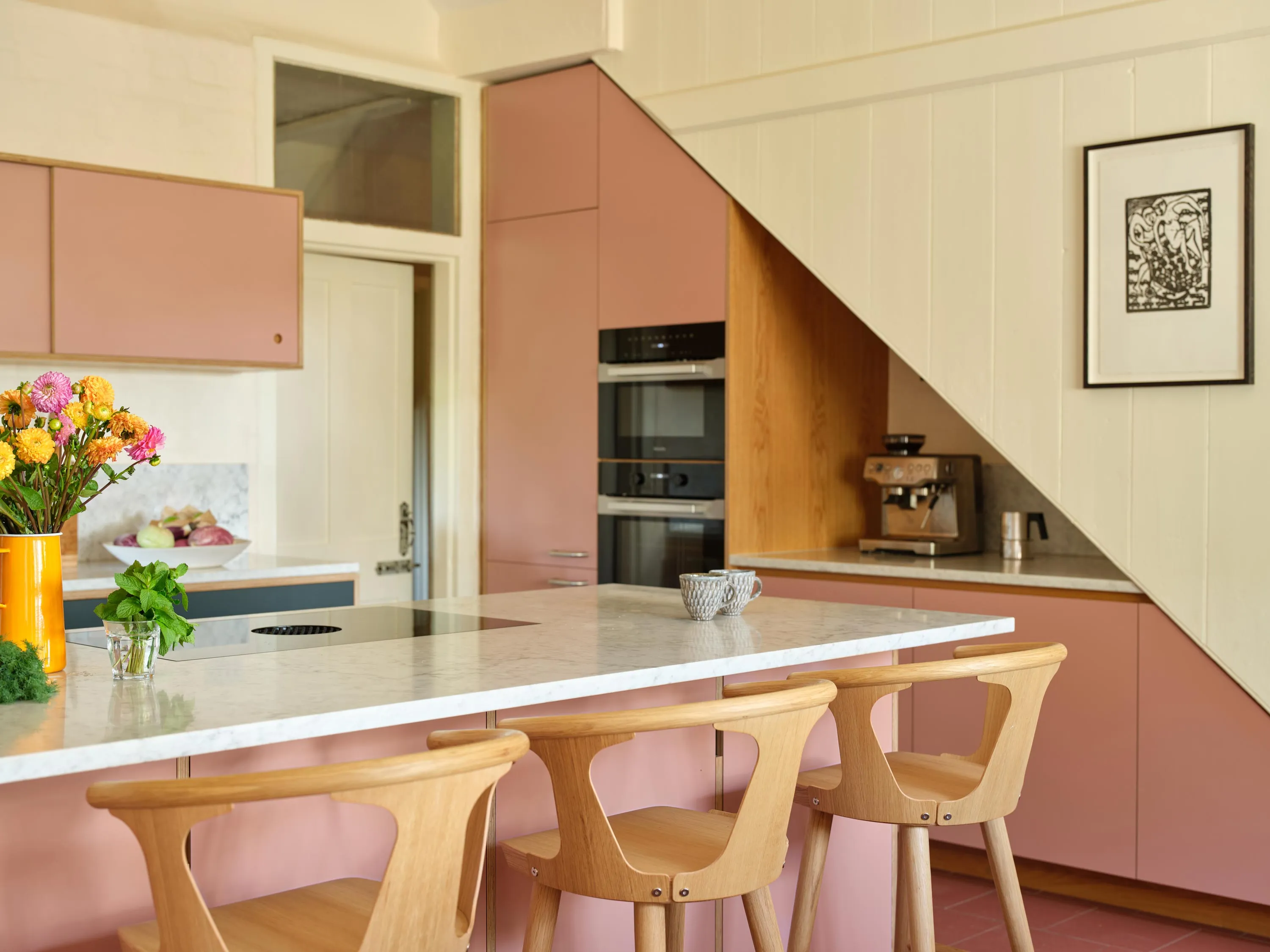 Modern kitchen with pink cabinets, white marble island with wooden stools, a coffee maker under the staircase, and a vase of colorful flowers.