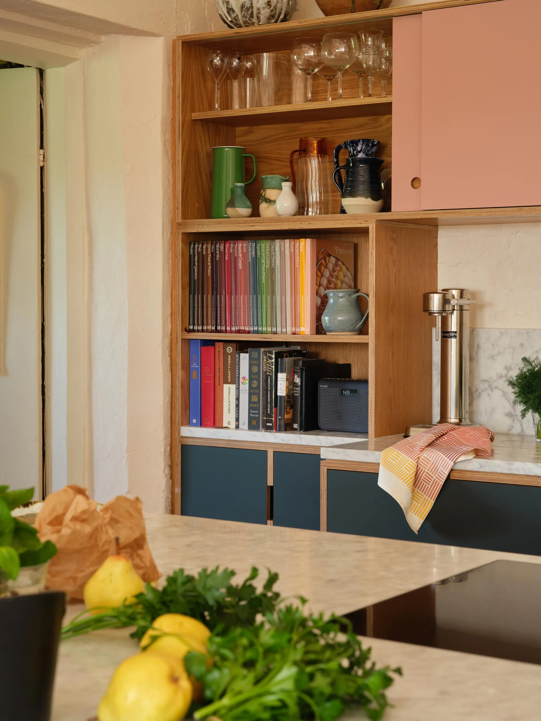 Wooden kitchen shelves with glassware, pitchers, cookbooks, and a radio, with a marble countertop holding a towel and herbs in the foreground.