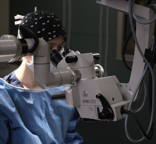 A woman in a blue surgical gown and head covering, wearing a bandana, is meticulously examining a microscope in a well-lit, professional setting.