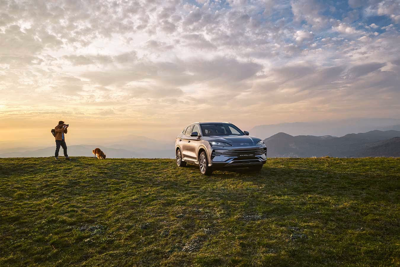 BYD SEAL U DM-i, Komplettansicht von 30° von vorne, Mann fotografiert mit Hund, Sonnenuntergang und Horizont im Hintergrund