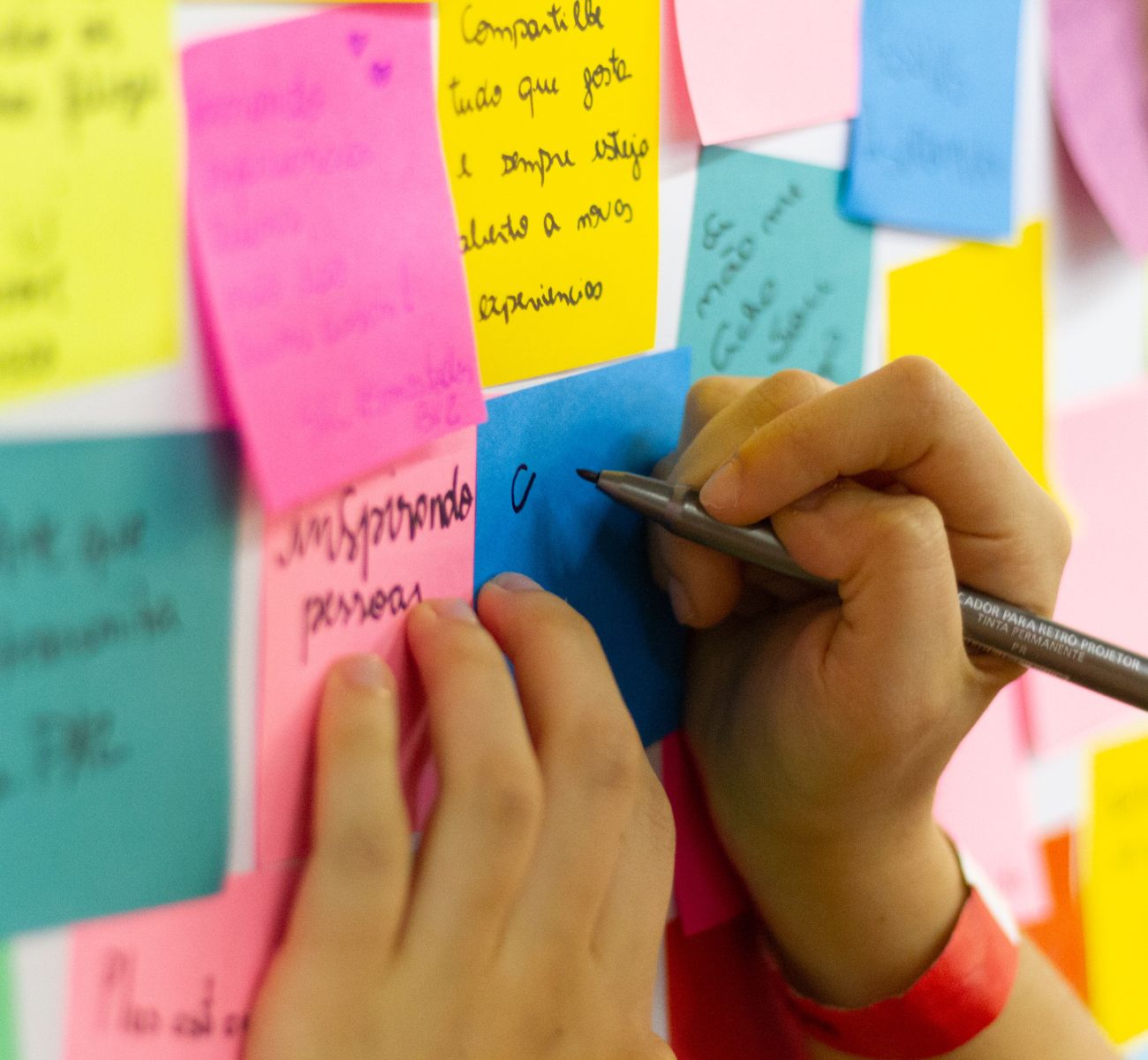 close up of a person's hand writing on a pink sticky note attached to a wall of sticky notes