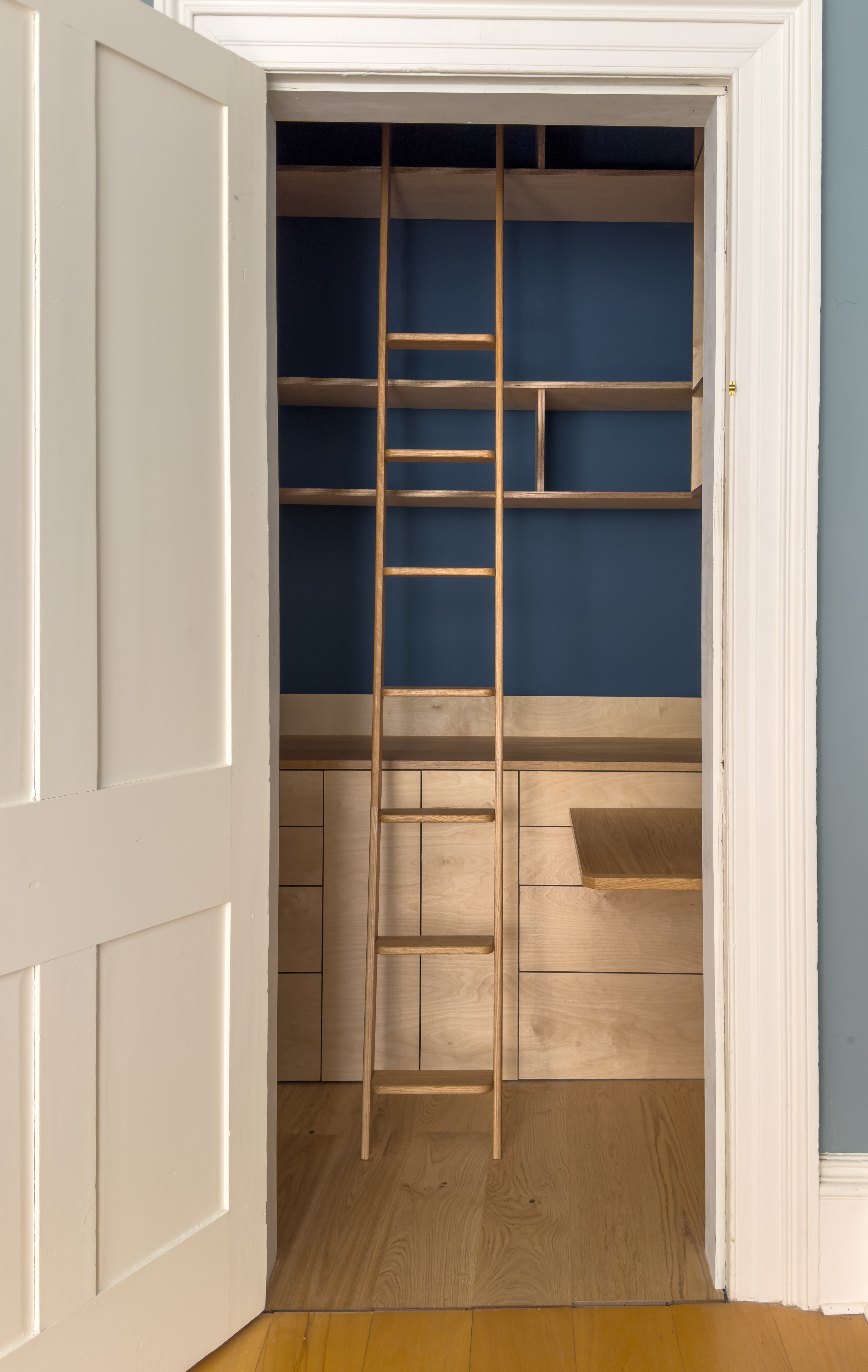 A photograph of the walk-in wardrobe, showing the rich tones of the wood against a handsome deep blue of the walls inside. The ladder to reach the higher shelves is visible.