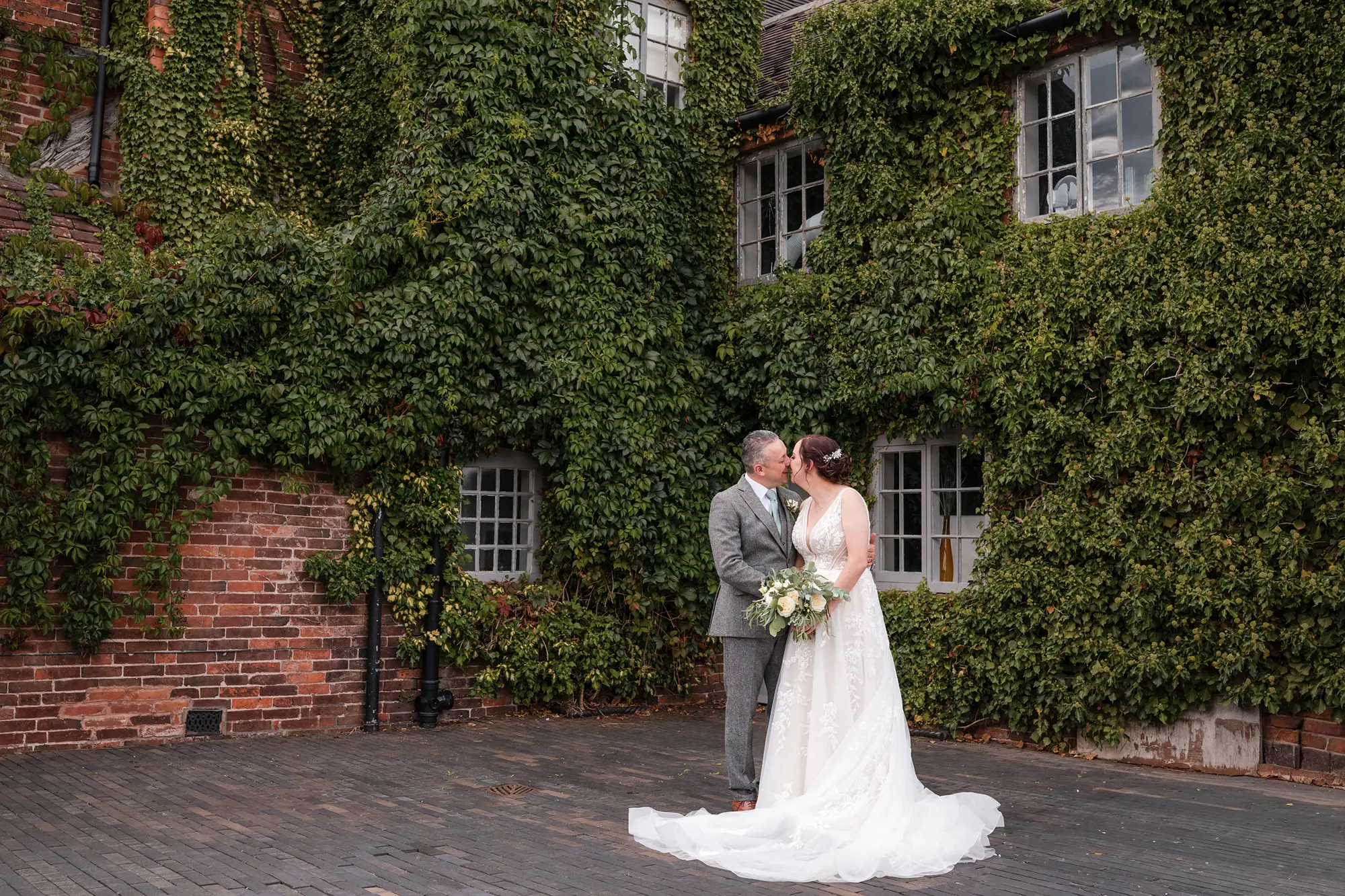 Newlyweds kissing beside the ivy-covered brick wall at Blackbrook Barns, Lichfield, Staffordshire