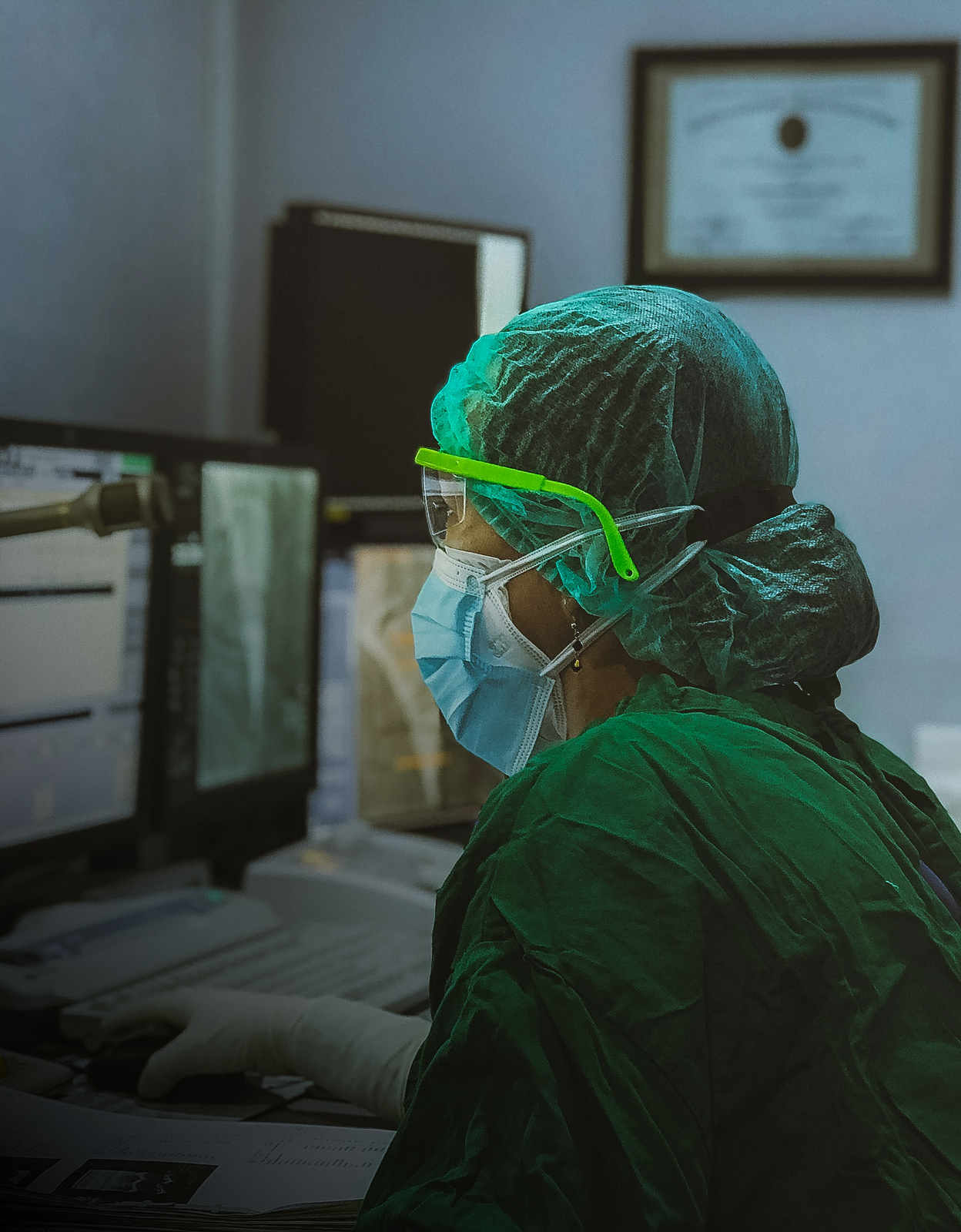 Healthcare worker in protective gear and face mask looking at computer monitors in a medical setting.
