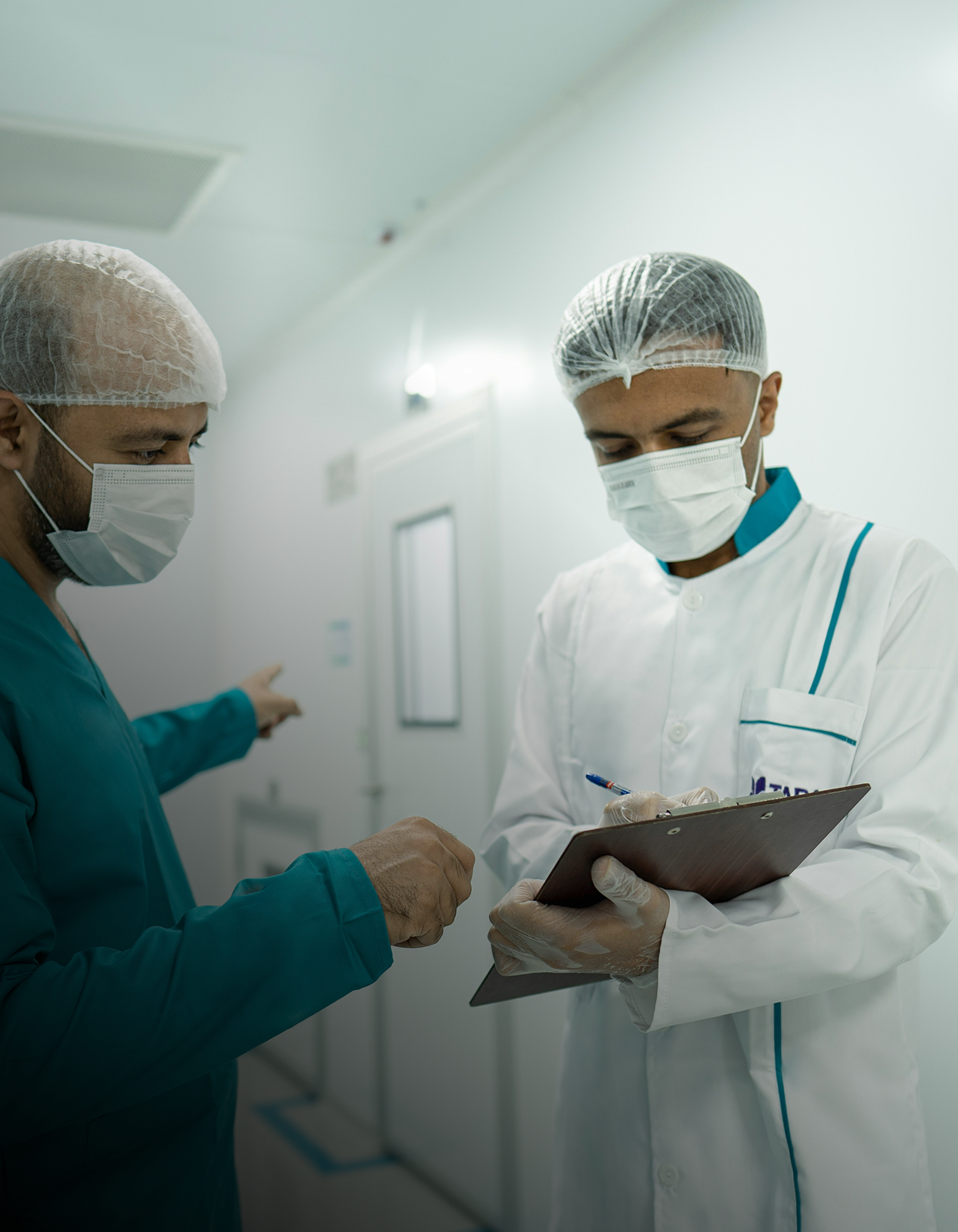 Two healthcare professionals wearing surgical masks and hair nets, one in blue scrubs pointing, the other in a white coat writing on a clipboard.