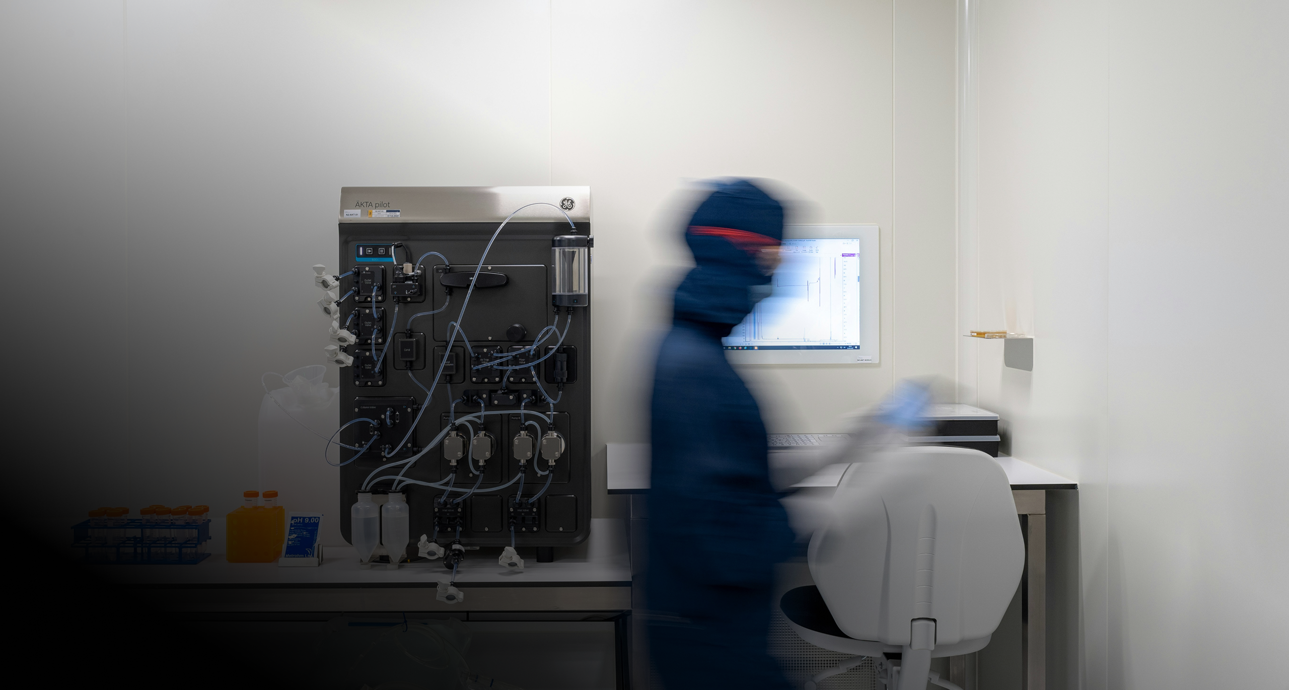 Blurry figure in protective lab clothing working near a chromatography machine and a computer in a clean laboratory.