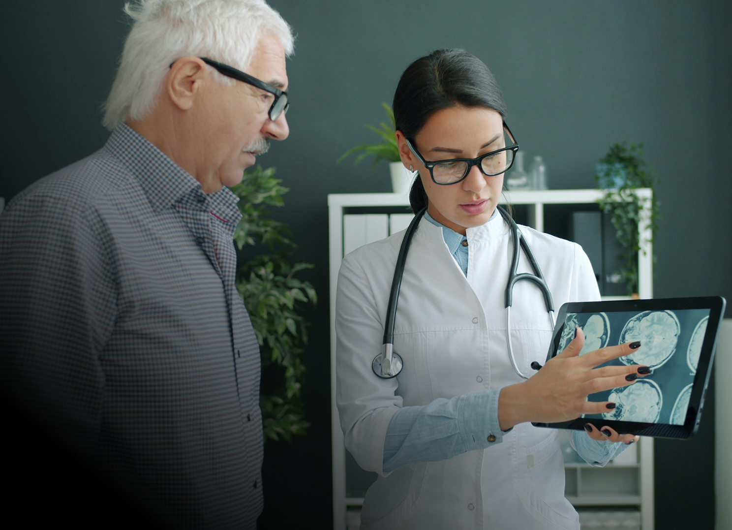 Female doctor showing brain scan images on a tablet to an older male patient in an office.