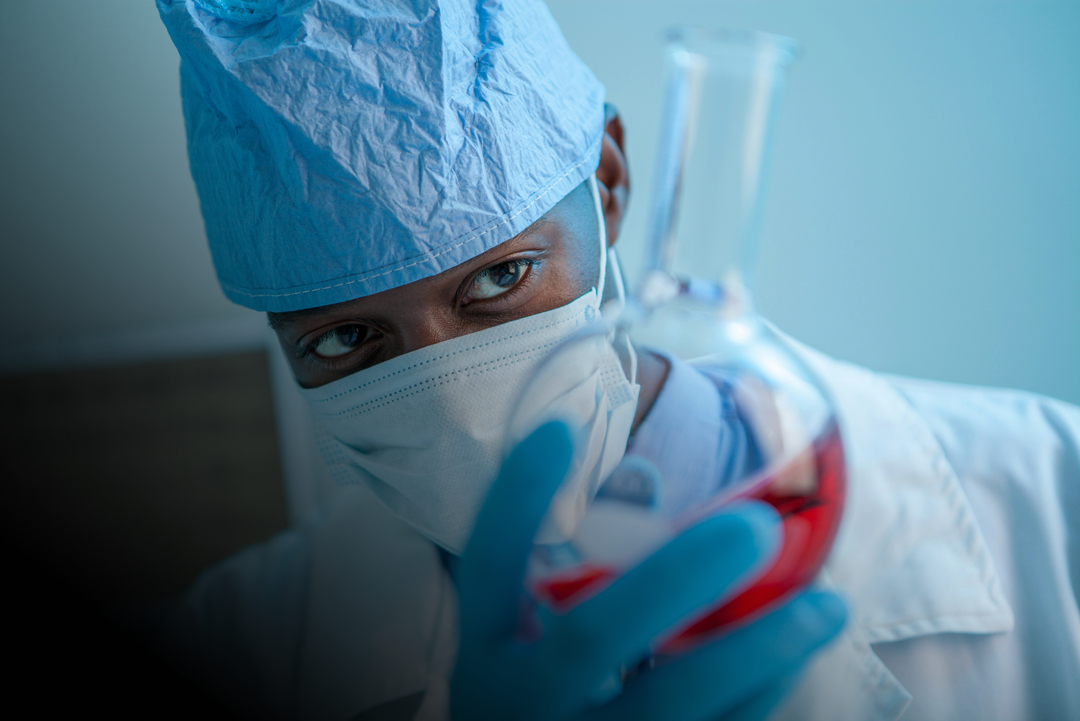 Scientist wearing blue gloves, a surgical cap, and mask examining a flask with red liquid.