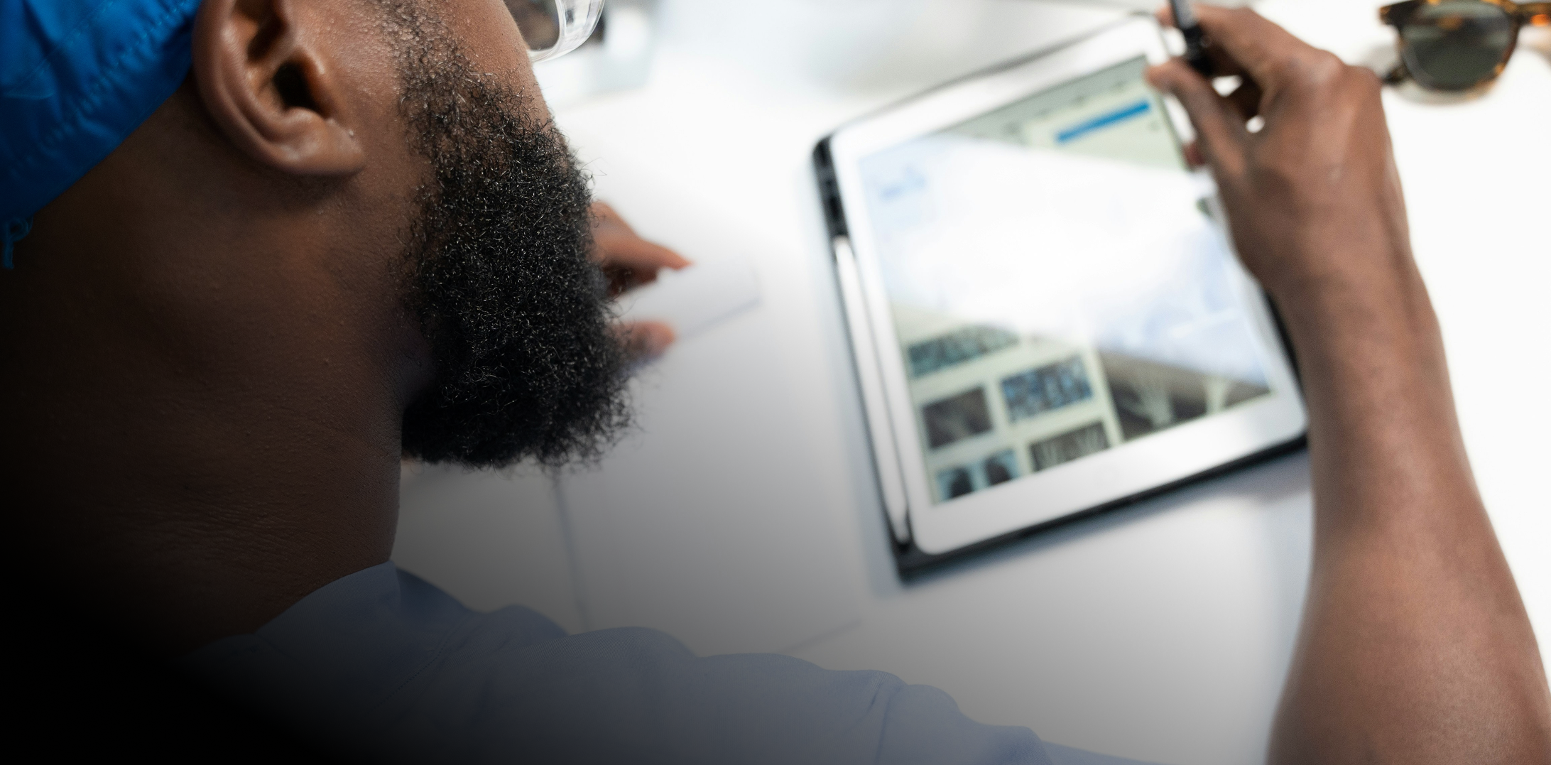 Close-up of a bearded man wearing glasses and a blue cap, using a digital tablet at a white desk.