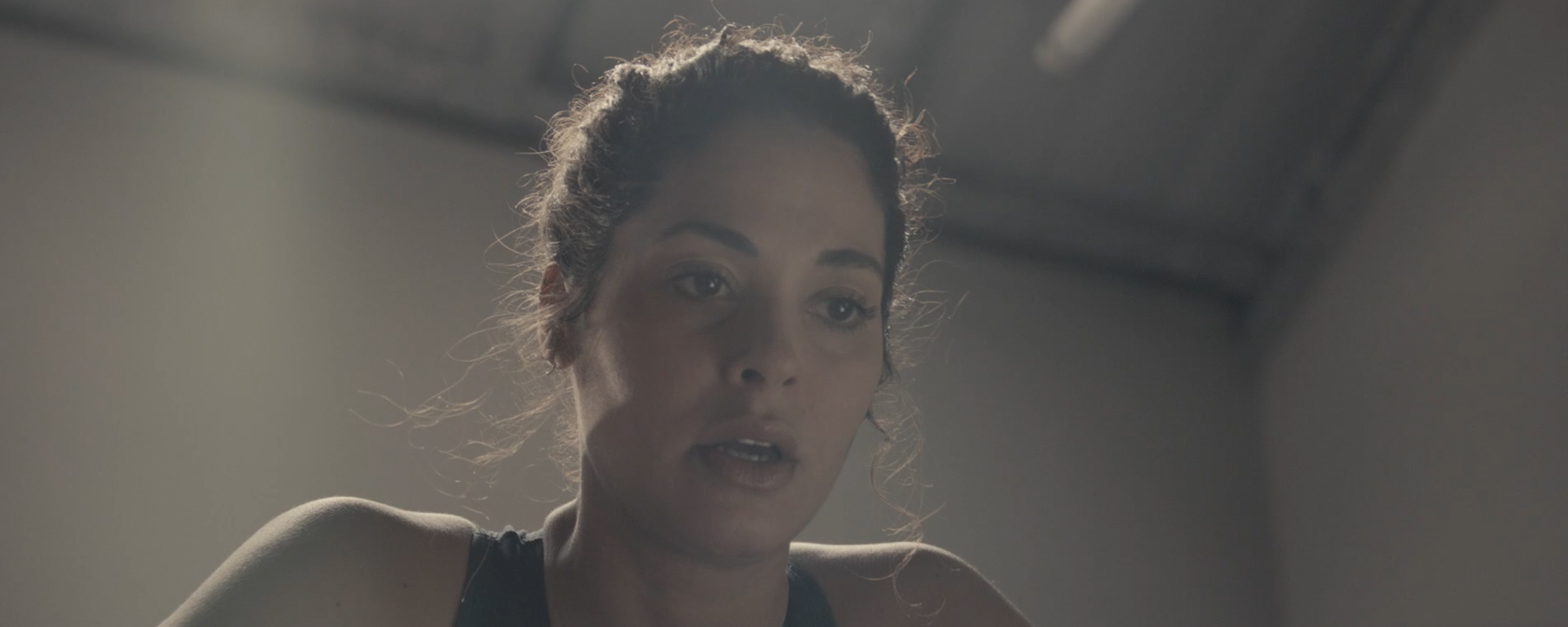 Femme à cheveux bouclés attachés, regardant vers le bas dans une pièce éclairée avec un plafond visible.