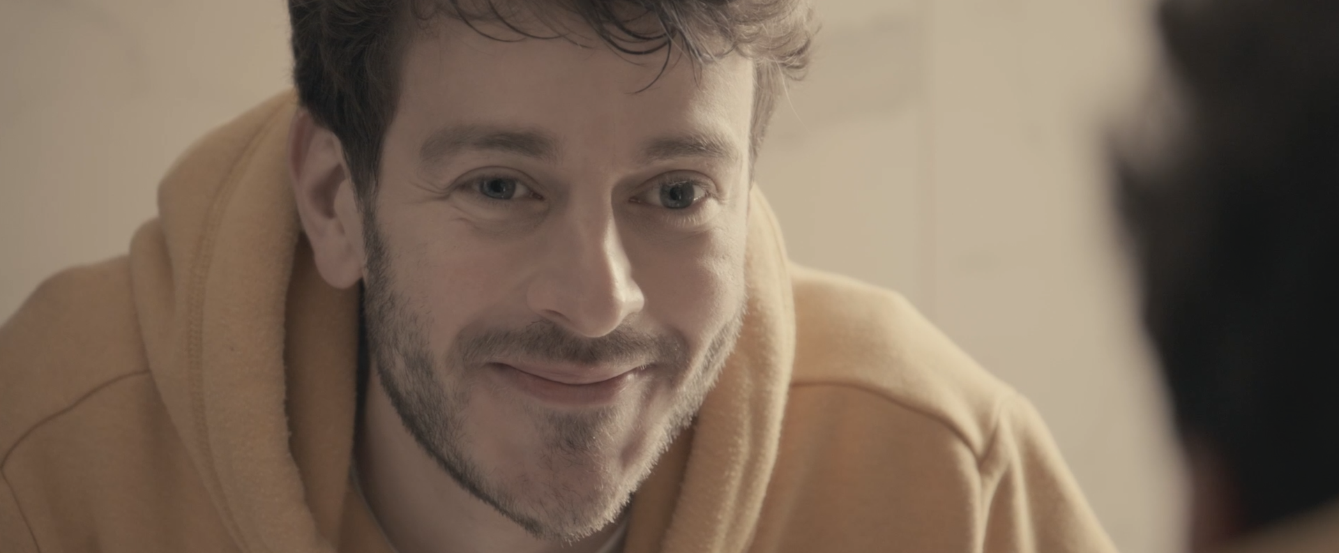 Jeune homme souriant portant un sweat à capuche beige, face à une personne floue au premier plan.
