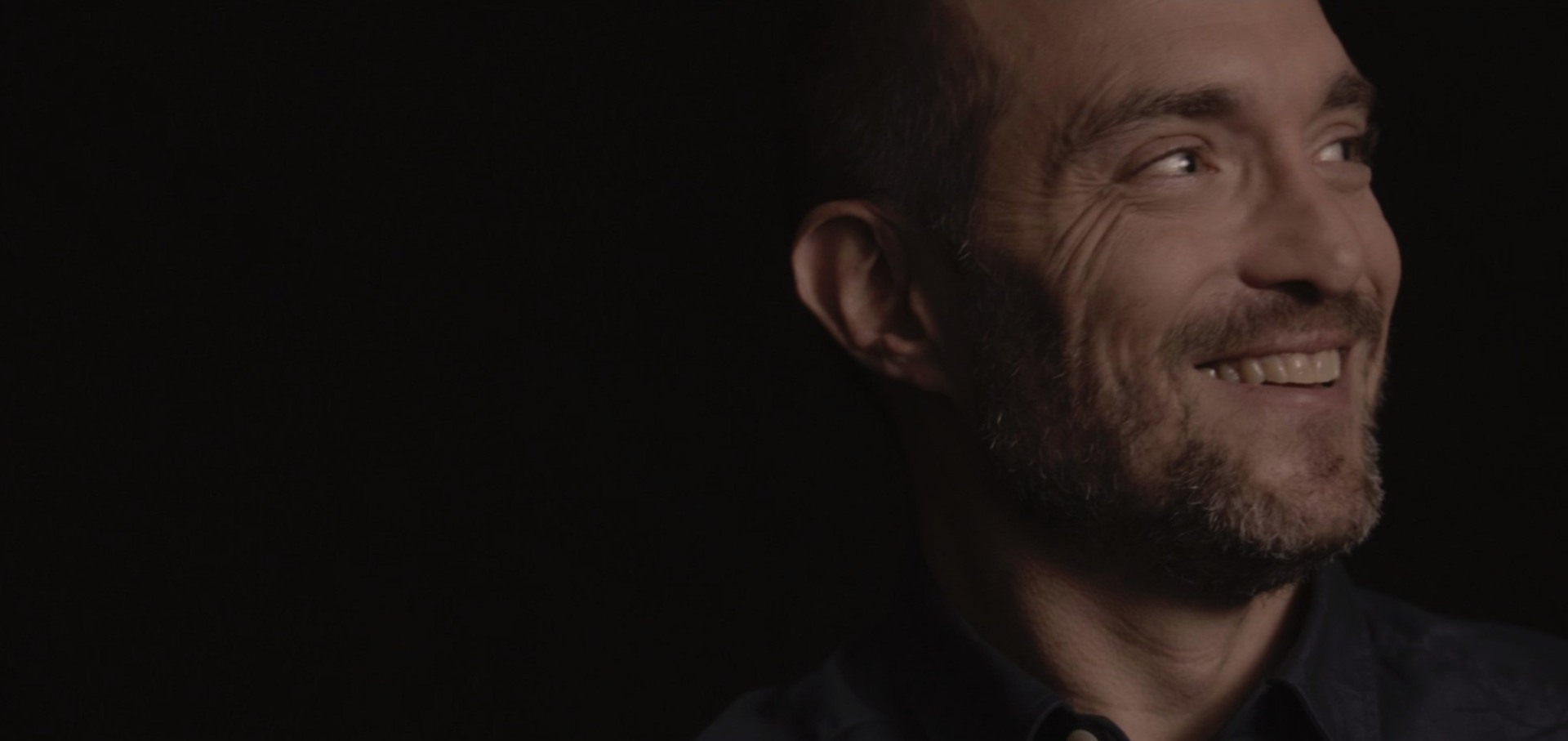 Portrait d'un homme souriant regardant vers la droite sur un fond sombre.