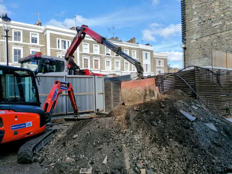 Construction site with an orange excavator moving a pile of soil in an urban area with residential buildings in the background.
