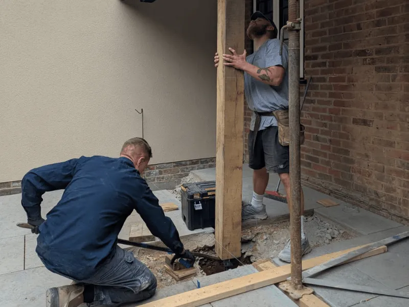 Two construction workers installing a wooden post on a cement floor near a brick wall, one worker holding the post upright and the other kneeling to work on the base.