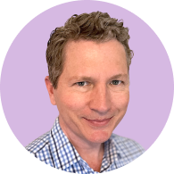 Smiling man with short curly hair wearing a blue and white checkered shirt on a light purple background.