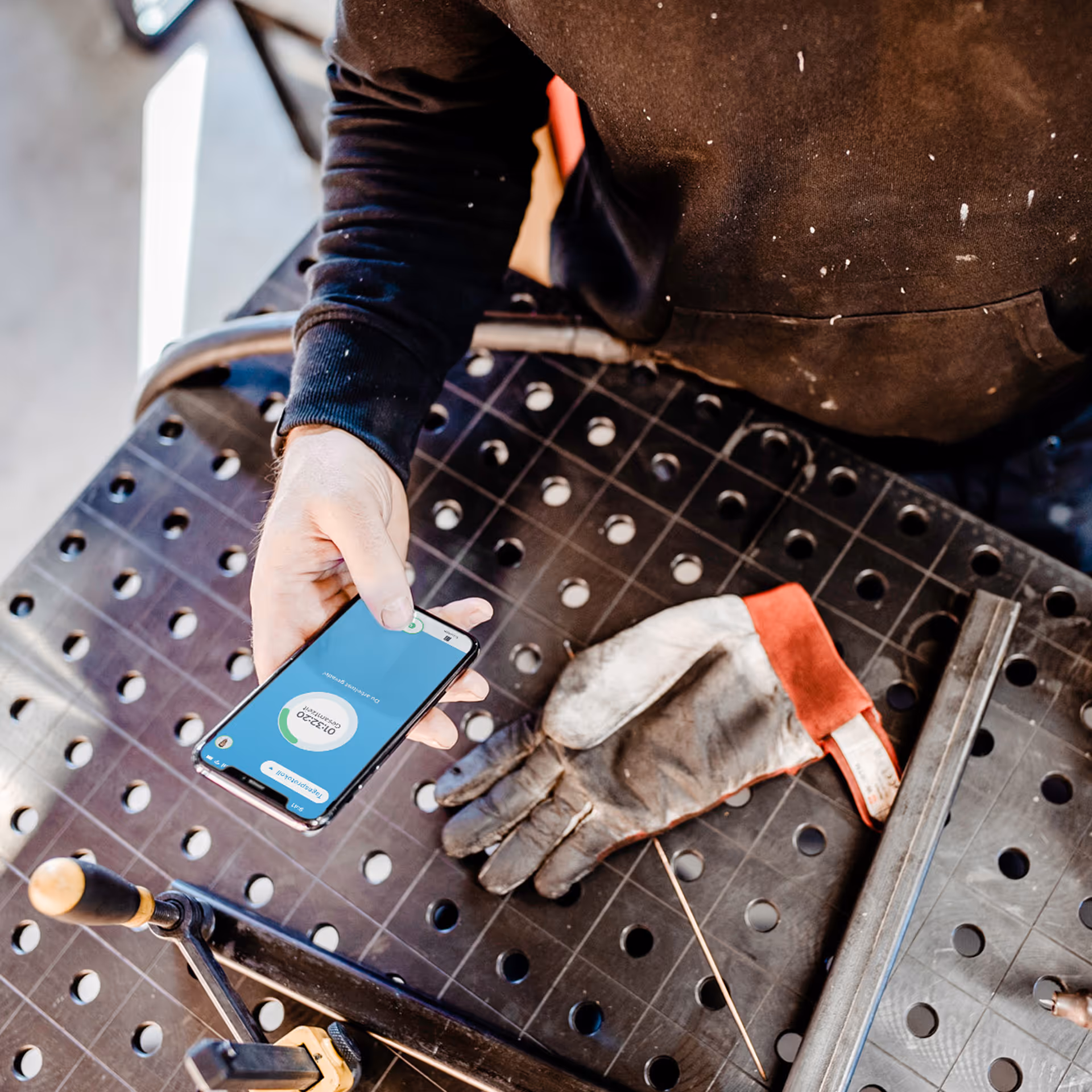Person hält Smartphone mit Anzeige auf Werkbank neben schmutzigem Arbeitshandschuh.