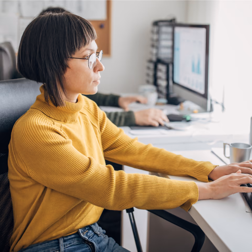 Frau mit Brille und gelbem Pullover arbeitet an einem Computer in einem Büro.