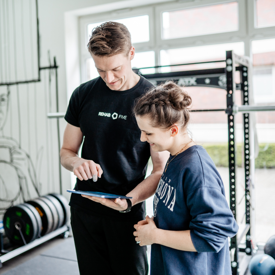 Trainer zeigt einer Frau im Fitnessstudio etwas auf einem Tablet. Beide lächeln und stehen nah beieinander.