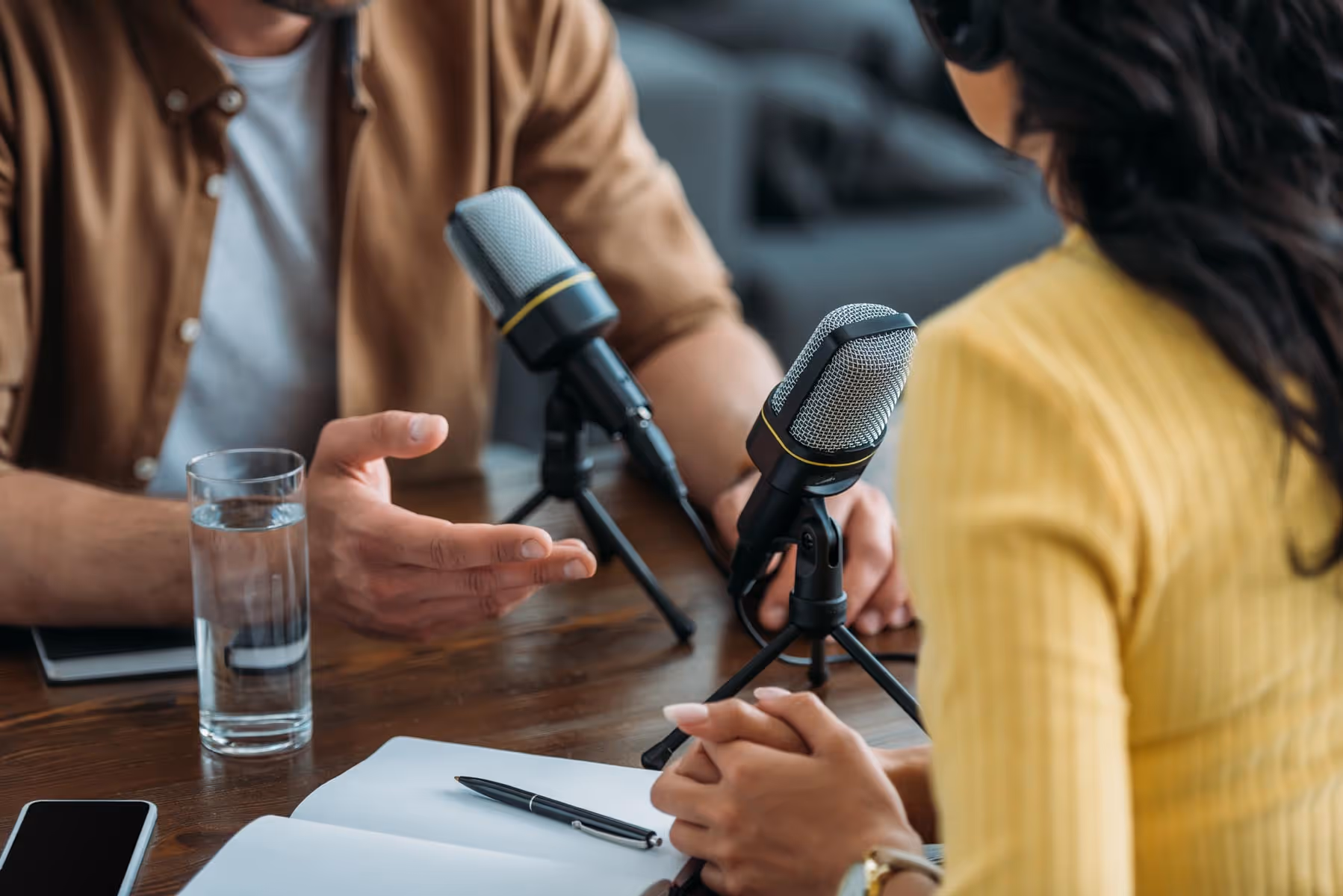 To personer som har en podcast-samtale med mikrofoner, notatbok, penn, glass vann og mobiltelefon på et trebord.