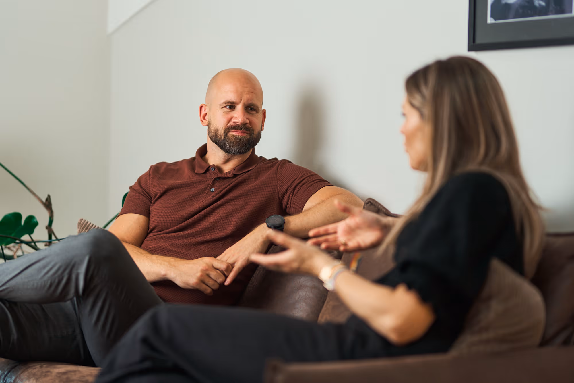 En mann i brun skjorte lytter oppmerksomt til en kvinne som snakker mens de sitter i en sofa.
