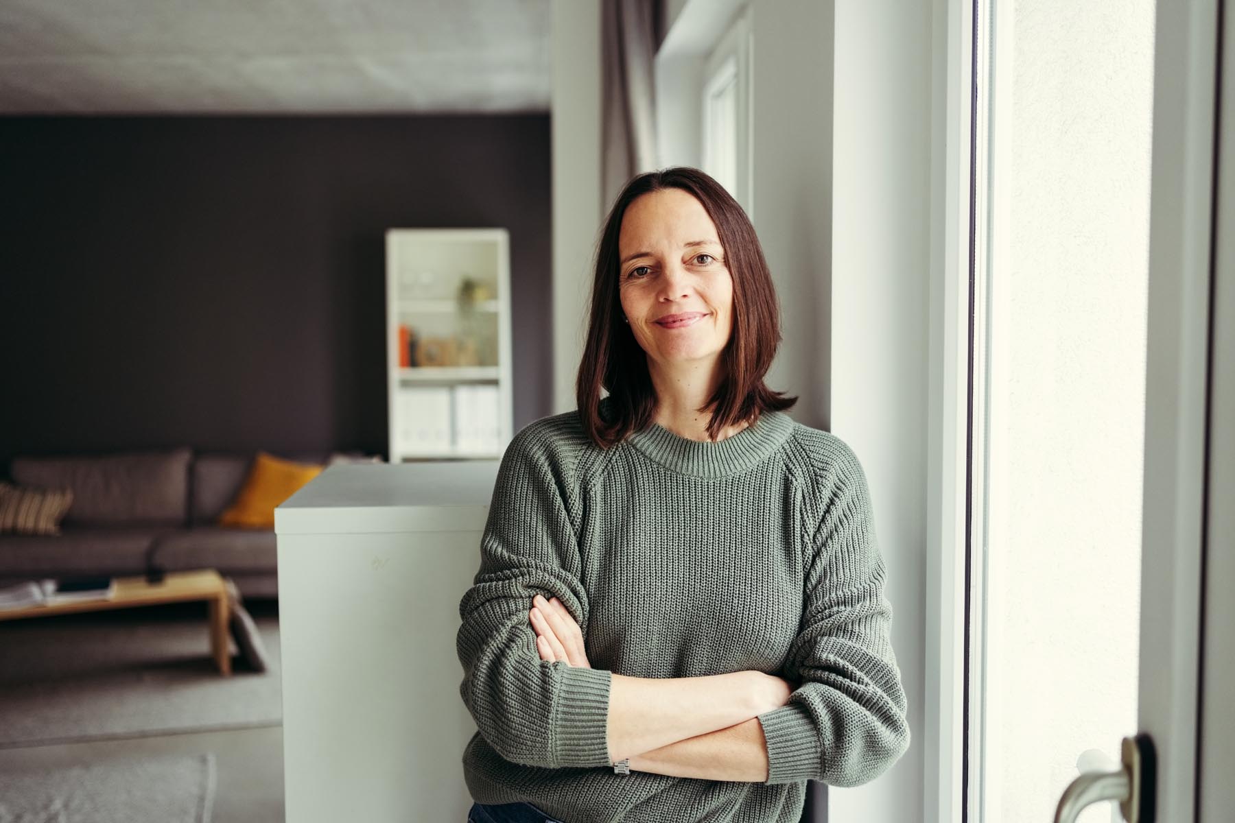 En kvinne som smiler til kameraet mens hun lener seg inntil veggen i sin egen stue