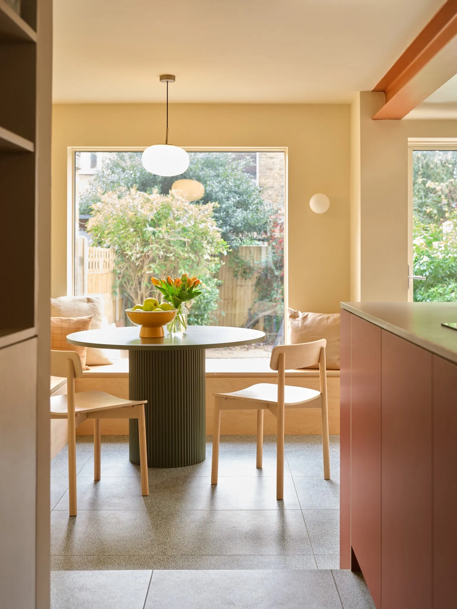 Dining area connected to the kitchen with bespoke plywood bench seating, pendant and large window facing the garden.