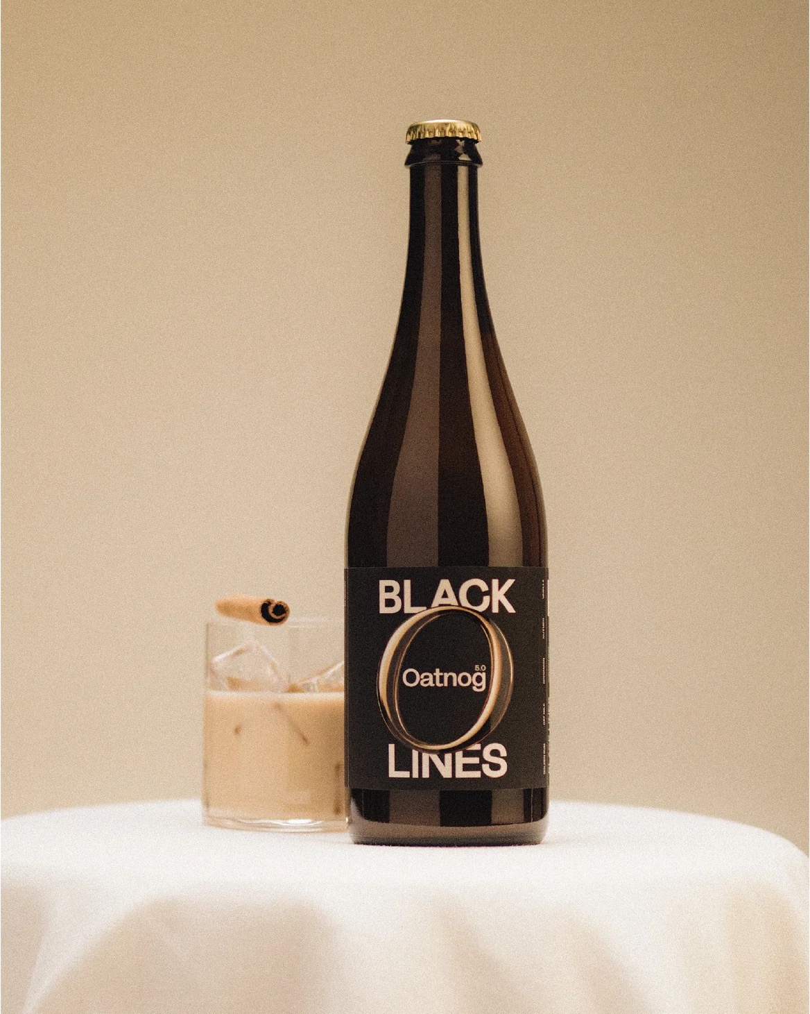 Bottle of Black Lines Oatnog with a glass of oat-based eggnog on a white table.