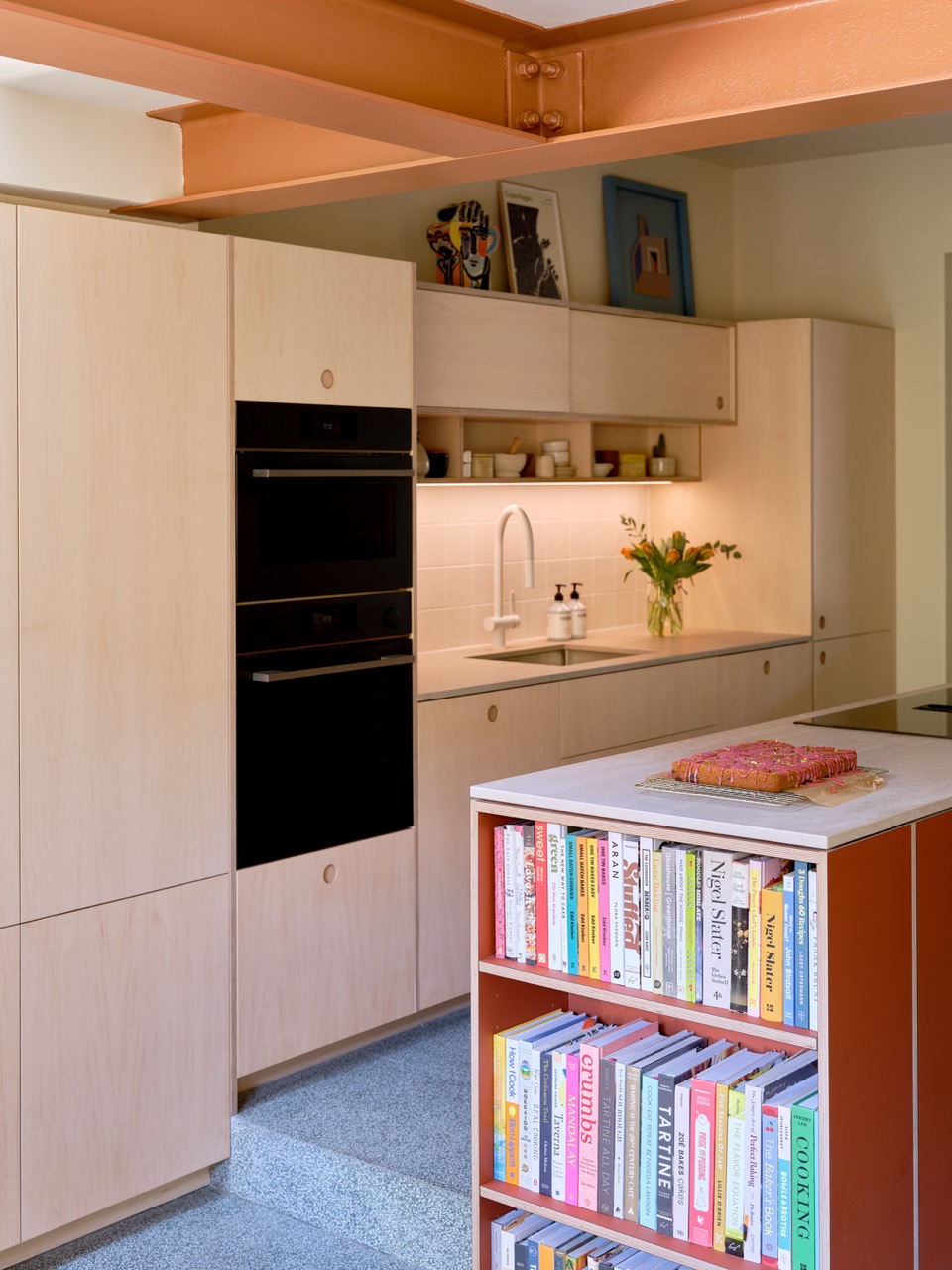Bespoke plywood kitchen for baker Edd Kimber in his Walthamstow home, designed by Uncommon Projects with maple veneer cabinetry, rust-coloured island with cookbook shelving, Caesarstone worktop and integrated ovens.