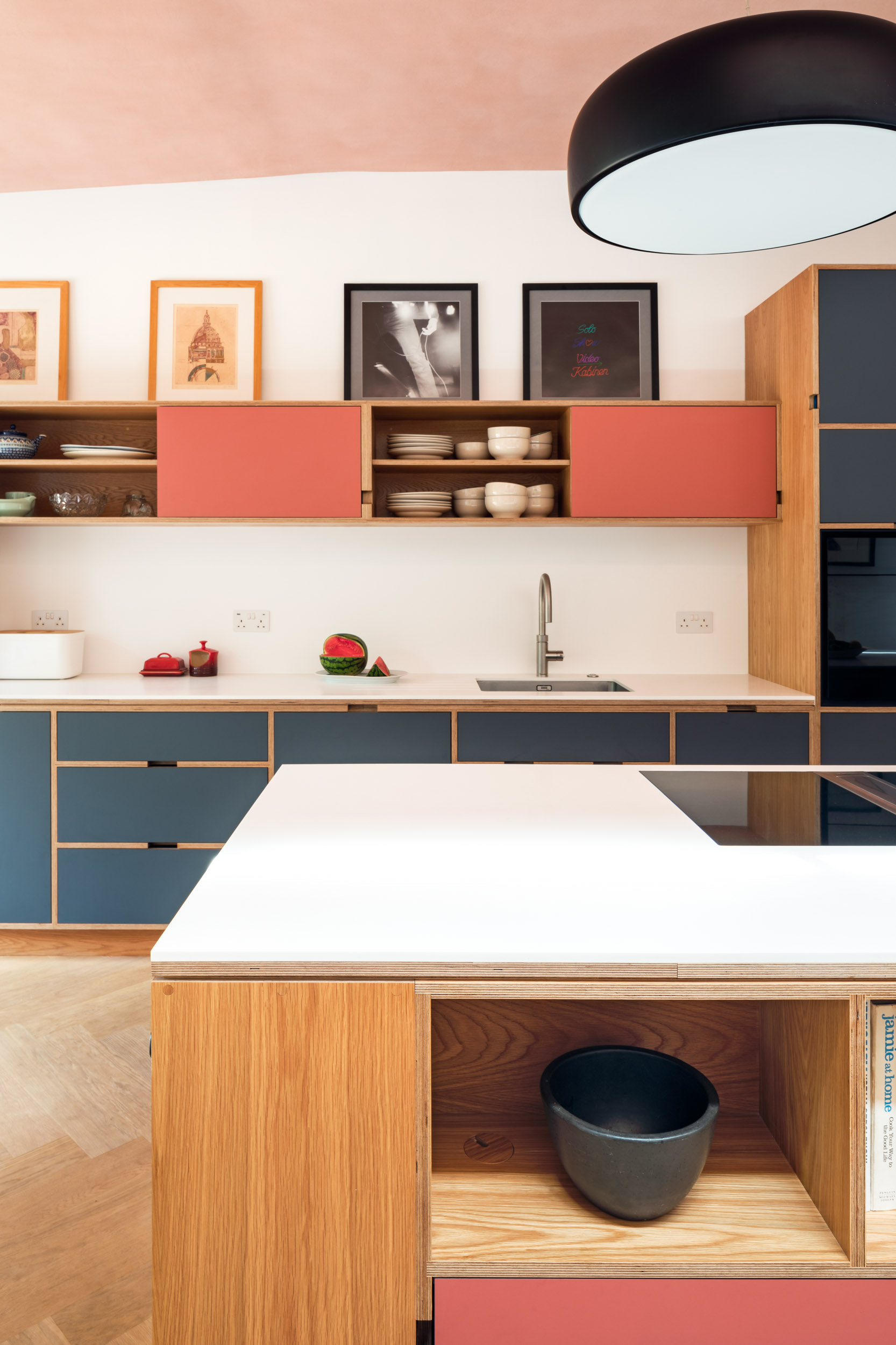 Close crop of oak plywood kitchen with pink and grey doors. and island