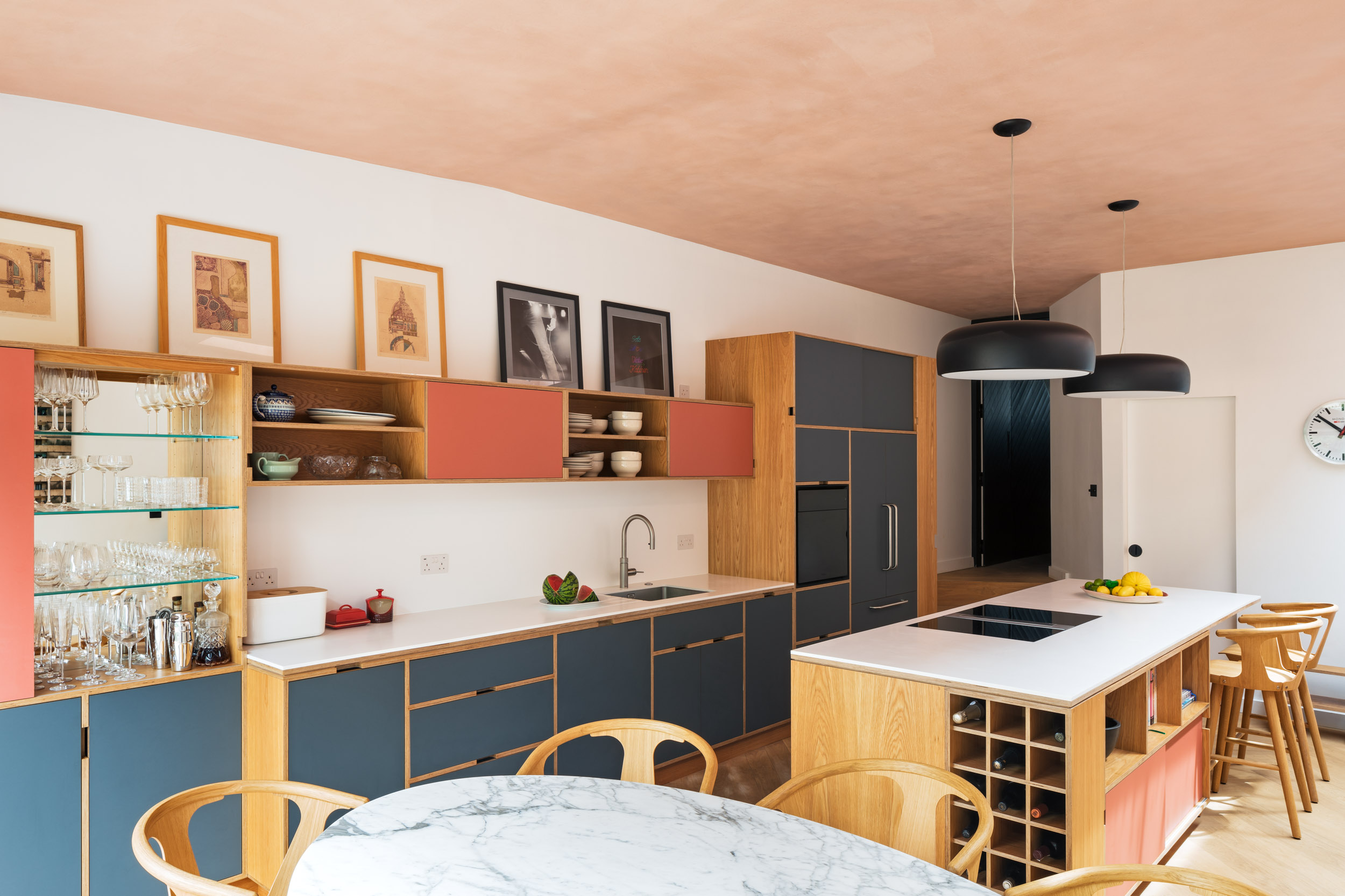 Bespoke oak plywood kitchen and island in Dulwich. Pictures on wall cabinetry. Art collection. Dusky pink doors with blue grey below.
