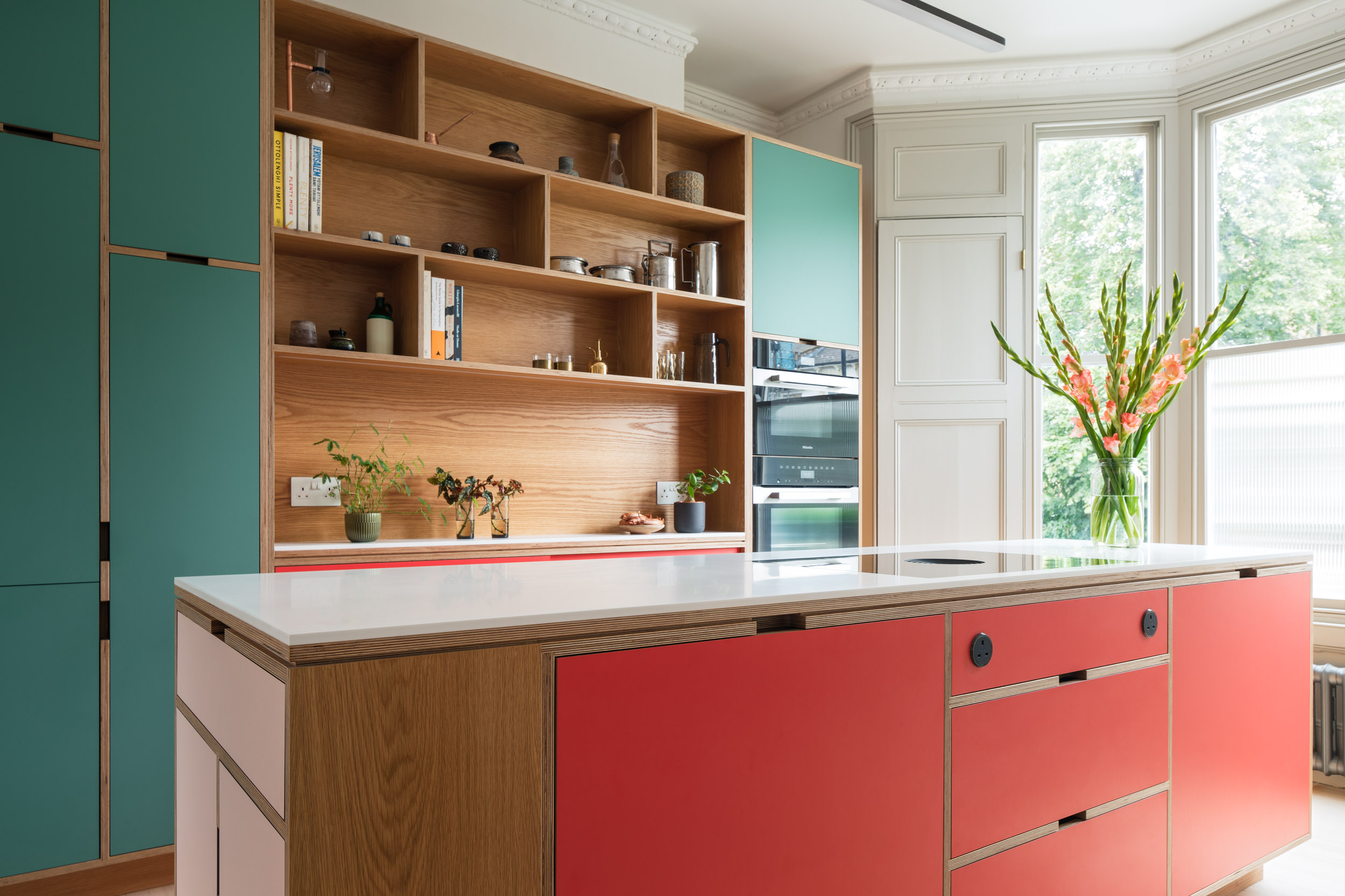 Bespoke multifunctional plywood open shelving, kitchen cabinetry and island. Pops of red, green and pink on the doors.