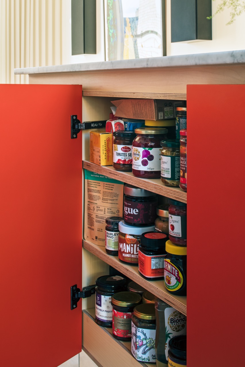 Open pantry cabinet door revealing plywood shelves stocked with jars, preserves and condiments