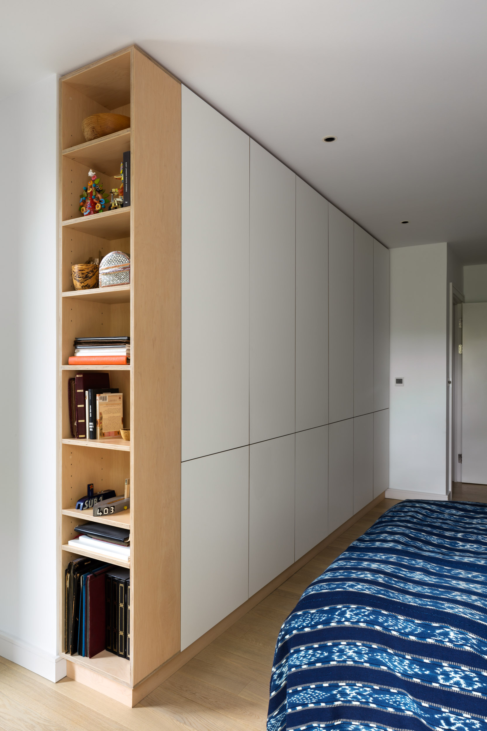Grey fitted wardrobe wall with an integrated birch plywood open bookcase in a bedroom.