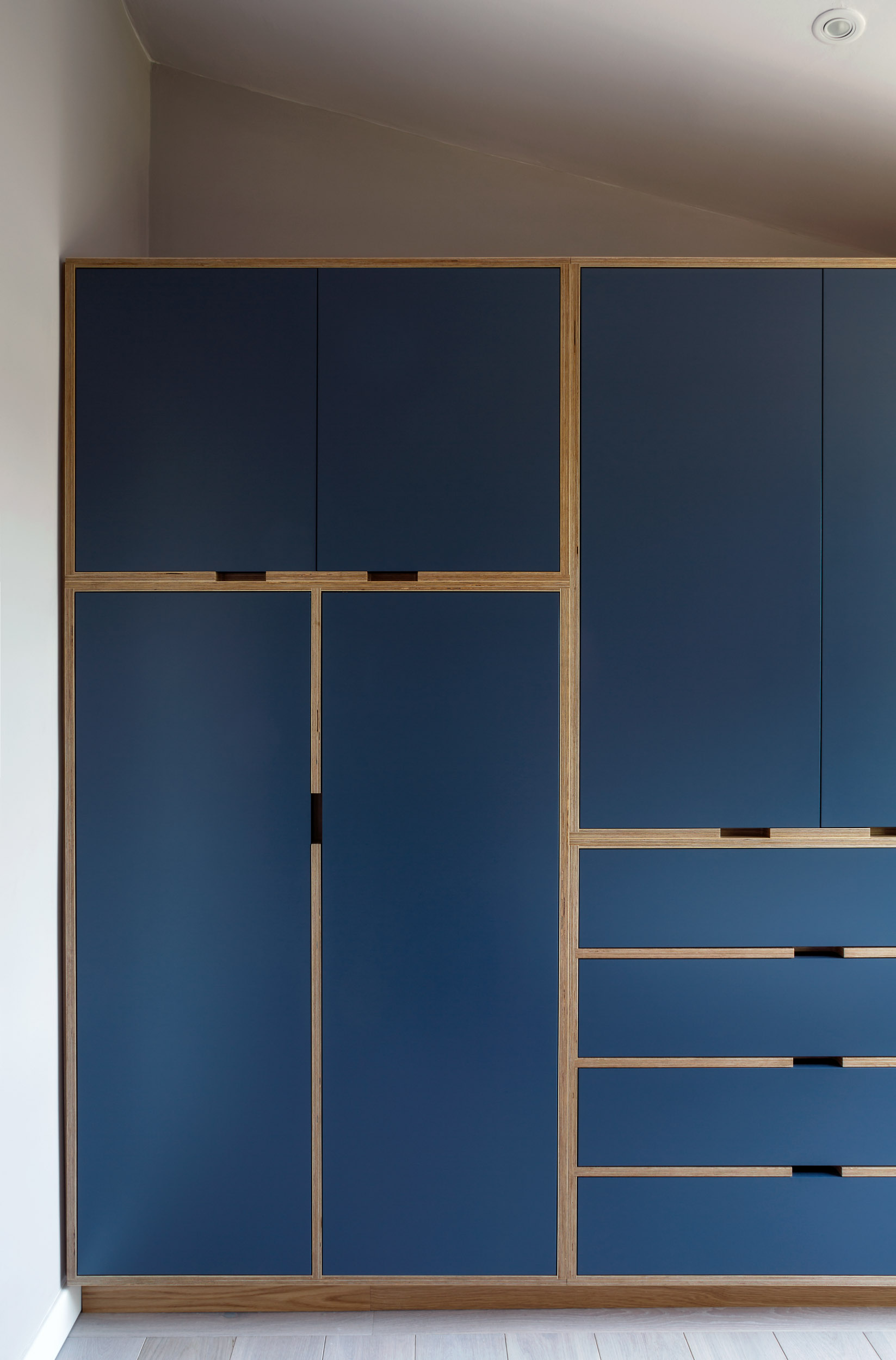 Floor-to-ceiling navy plywood fitted wardrobe with birch ply reveals, built under a sloping ceiling.