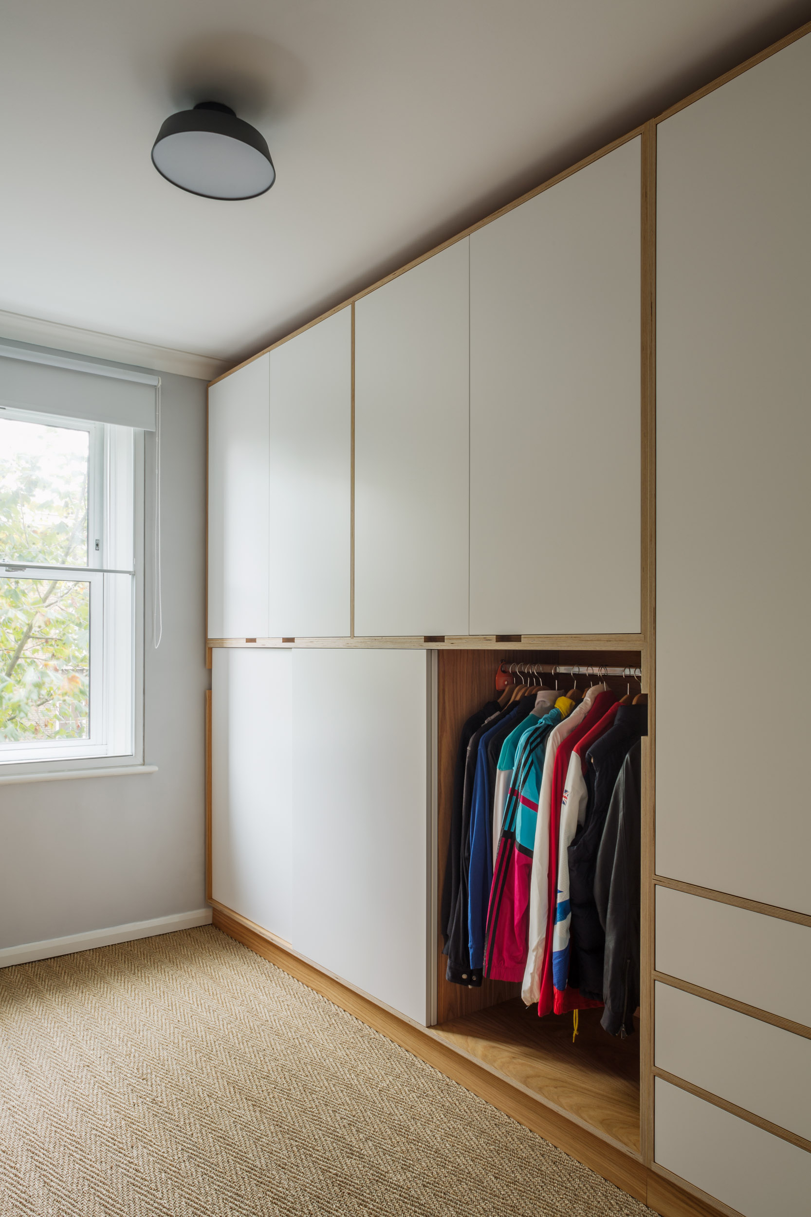 Cream plywood fitted wardrobe with an open bay of hanging space beside a bedroom window.