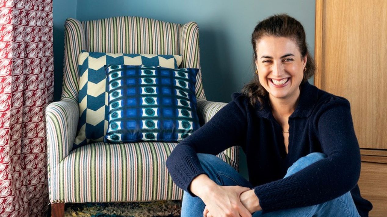 Interior designer Kate Guinness sitting on a colourful rag rug beside a striped armchair in a blue-walled period room