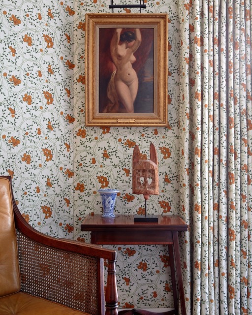 Rust floral wallpaper and matching curtains with a gilt-framed nude painting and a cane armchair in a period London apartment