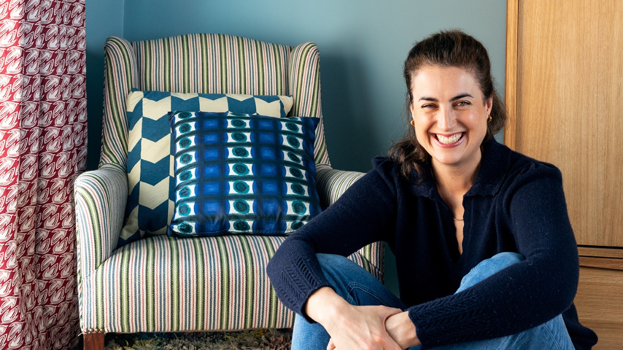 Interior designer Kate Guinness sitting on a colourful rag rug beside a striped armchair in a blue-walled period room