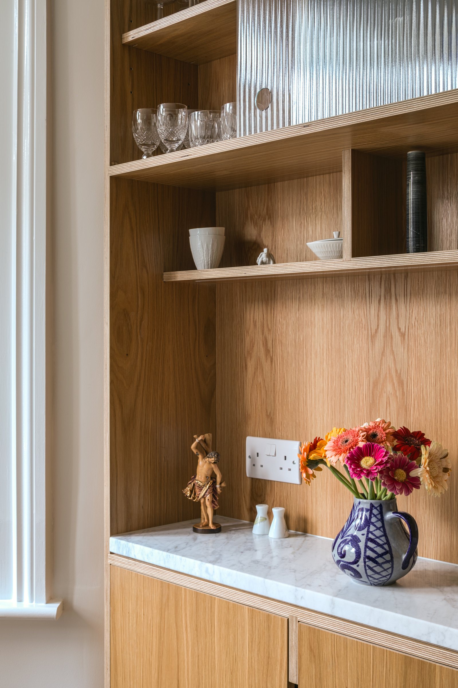 Oak veneer alcove cabinet with open shelves, fluted glass door and Carrara marble worktop holding a jug of gerberas