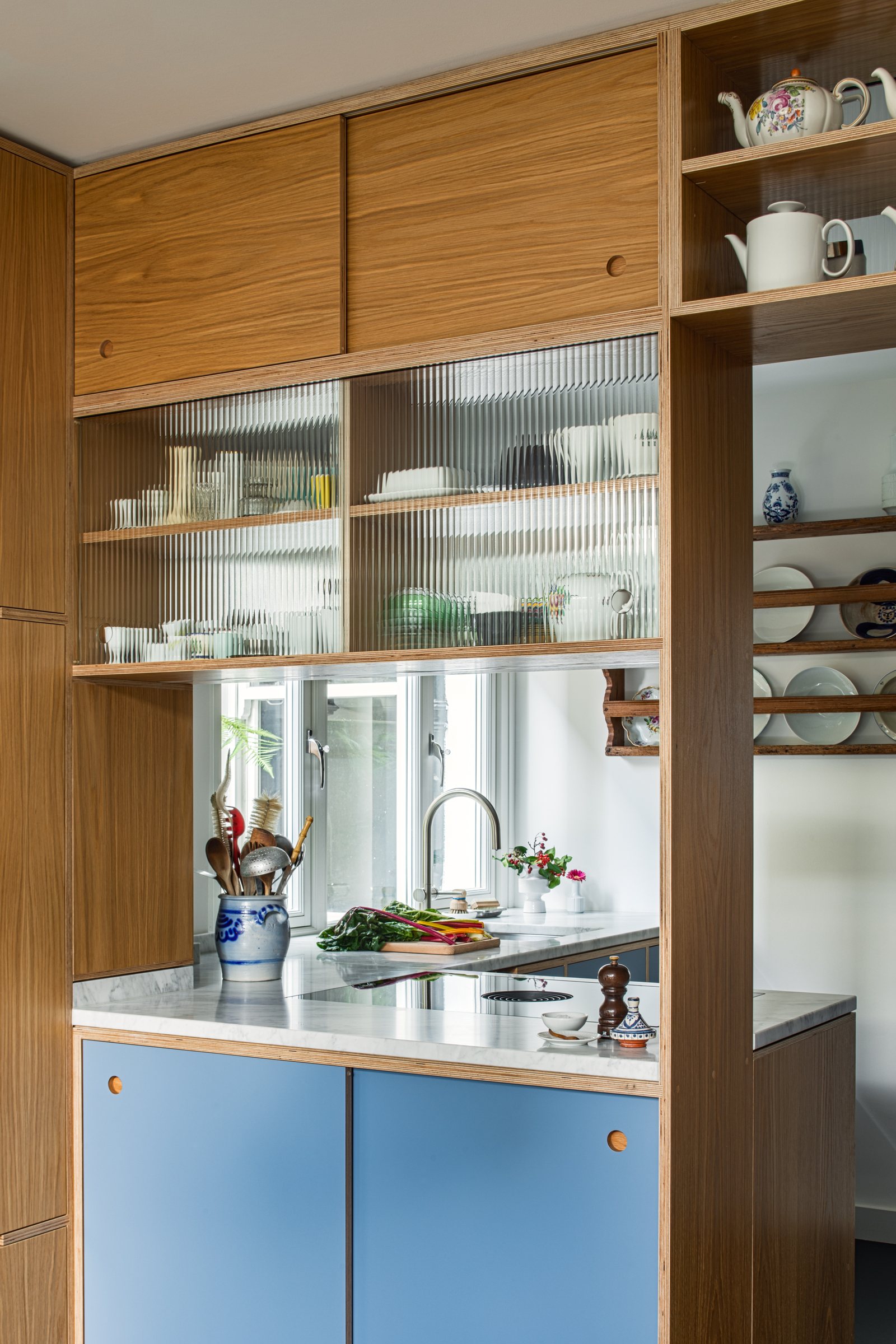 Oak and blue plywood screen with fluted glass display shelves and a hatch opening onto the kitchen beyond