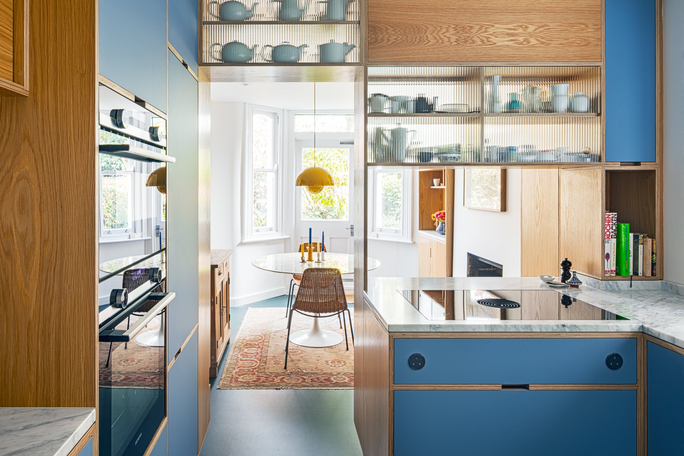 Galley kitchen with blue laminate units and Siemens ovens, looking through an oak screen to the dining room bay window