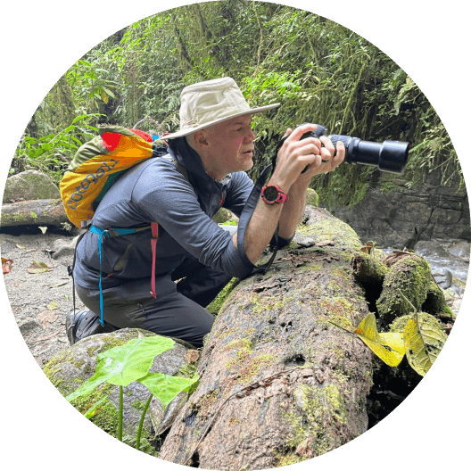 Photographer leaning behind a tree trunk with a camera