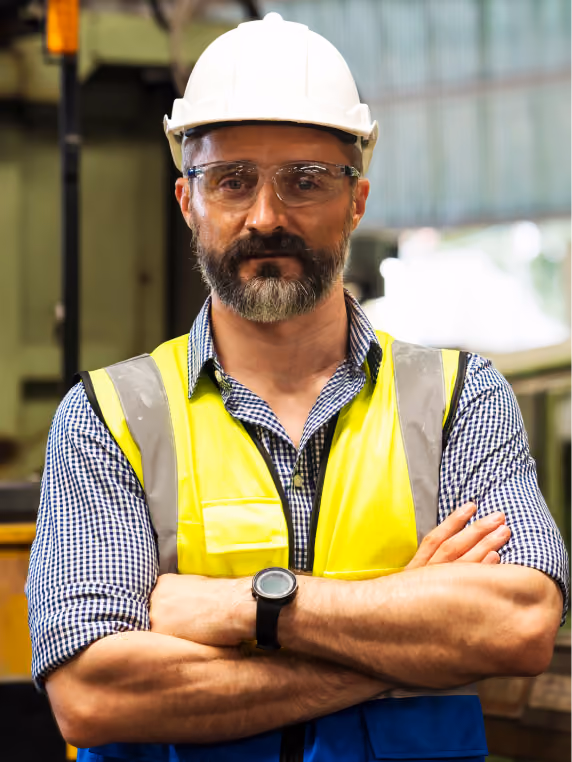 Male industrial worker wearing a white safety helmet, safety glasses, a yellow reflective vest, and a checkered shirt, standing with arms crossed in a factory setting.