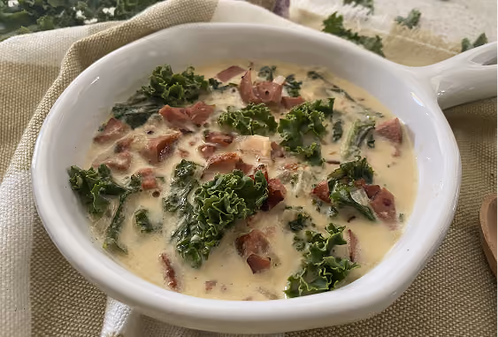 Creamy soup with chopped kale and pieces of bacon served in a white bowl with a handle on a beige cloth.
