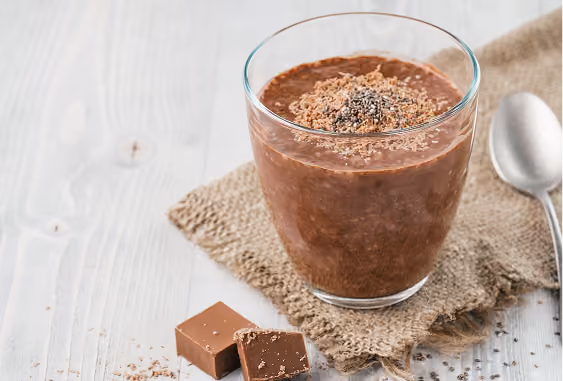 Glass of chocolate chia seed pudding topped with shredded chocolate and chia seeds on a burlap cloth next to two pieces of chocolate and a spoon on a light wooden surface.