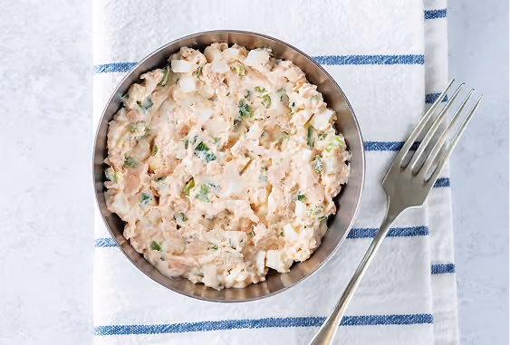 Bowl of creamy tuna salad with chopped eggs and herbs on a white and blue striped cloth with a fork beside it.